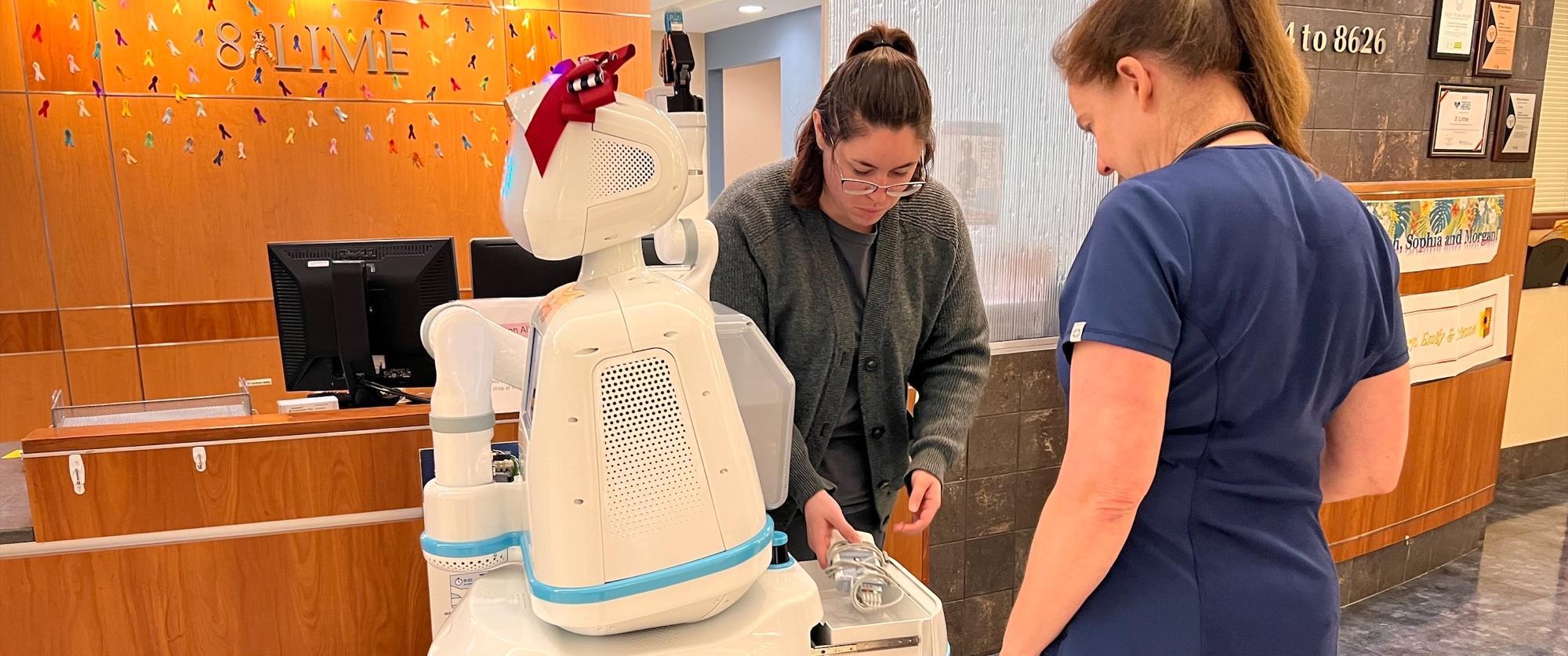  Two nurses interact with a human-sized white robot wearing a red bow atop her head