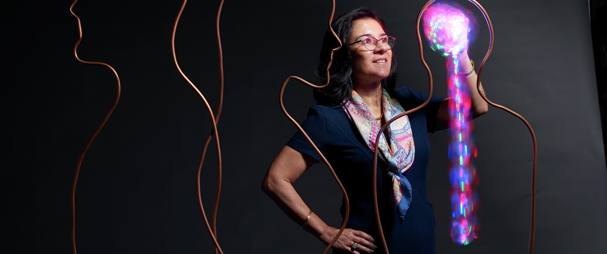 Maria Oquendo stands with one hand appearing enmeshed in a brain shape made of light, within and surrounded by abstract outlines of human silhouettes
