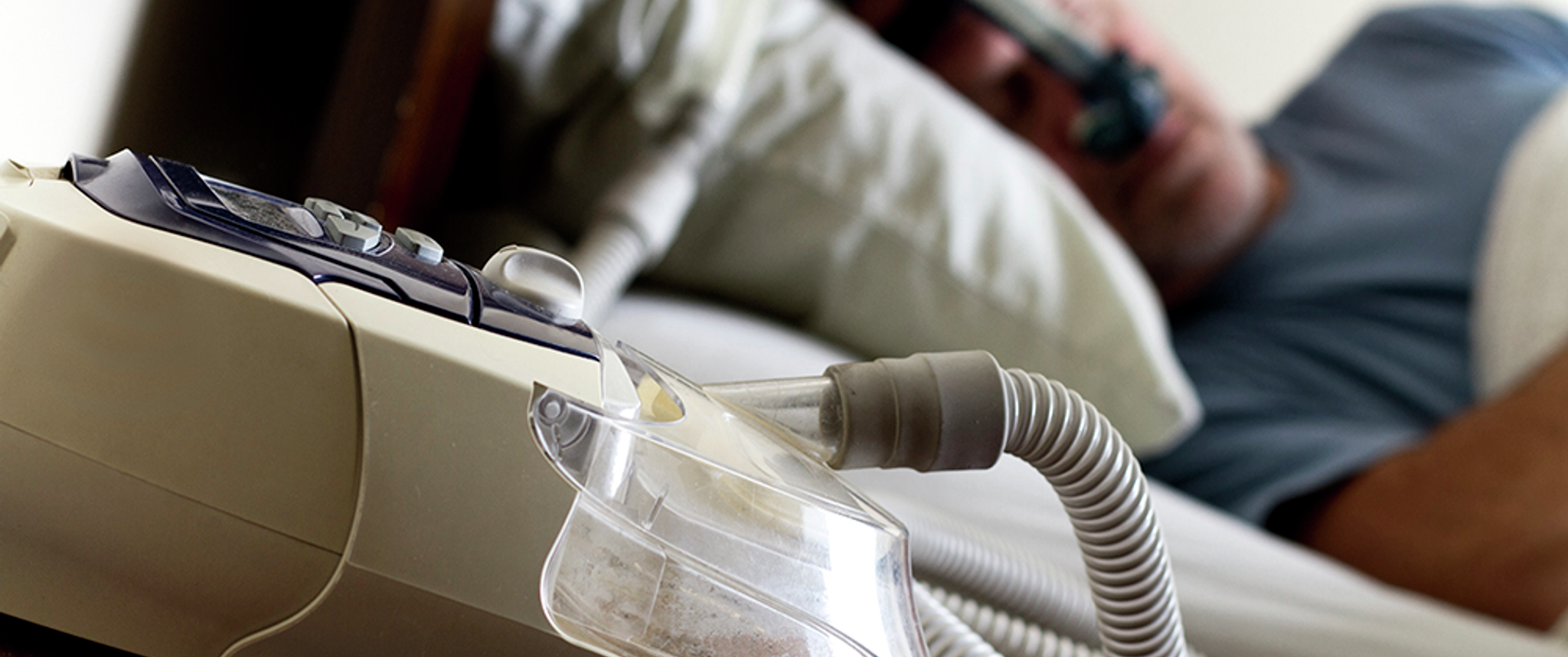 A man sleeps wearing a CPAP mask in the background; in the foreground a CPAP machine rests on a table