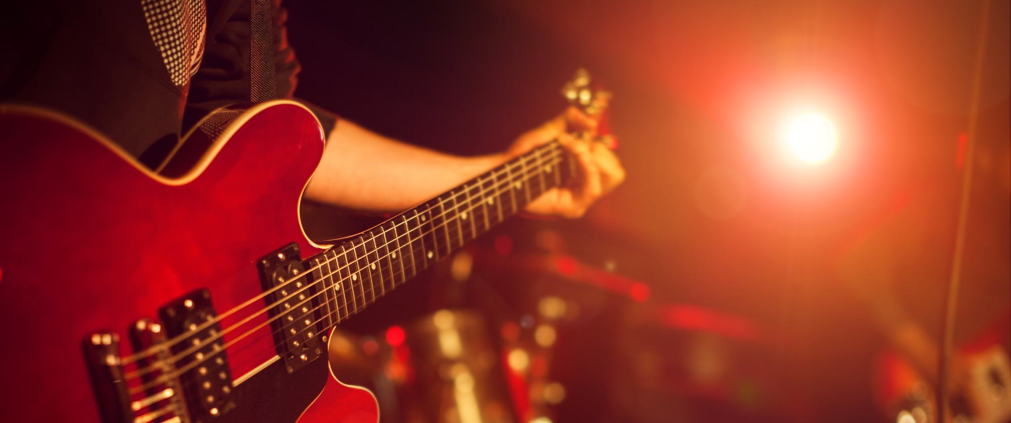 A guitarist playing a guitar on stage with a drum set in the background.