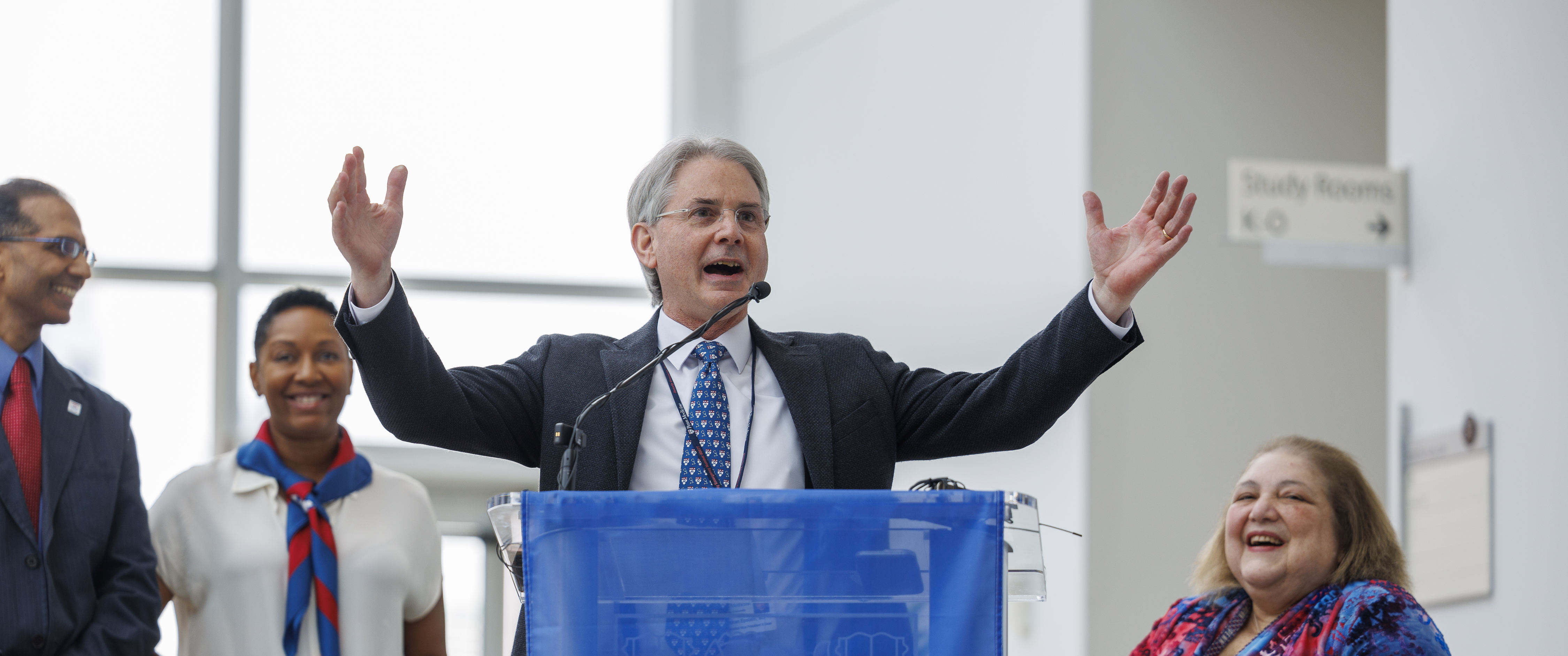 Jonathan Epstein, MD, stands behind a podium with arms raised in celebration while other PSOM leaders smile behind him.