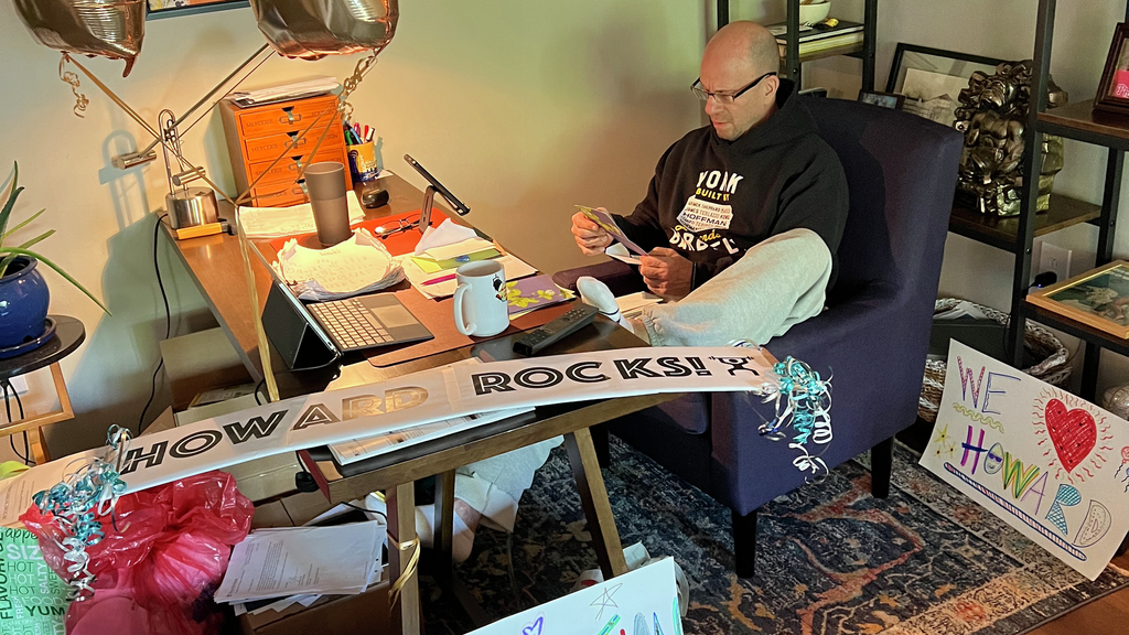 Howard Aaron sits behind his desk in his home office, surrounded by balloons and posters that display phrases like “Super Strong” and “Inspiration.”