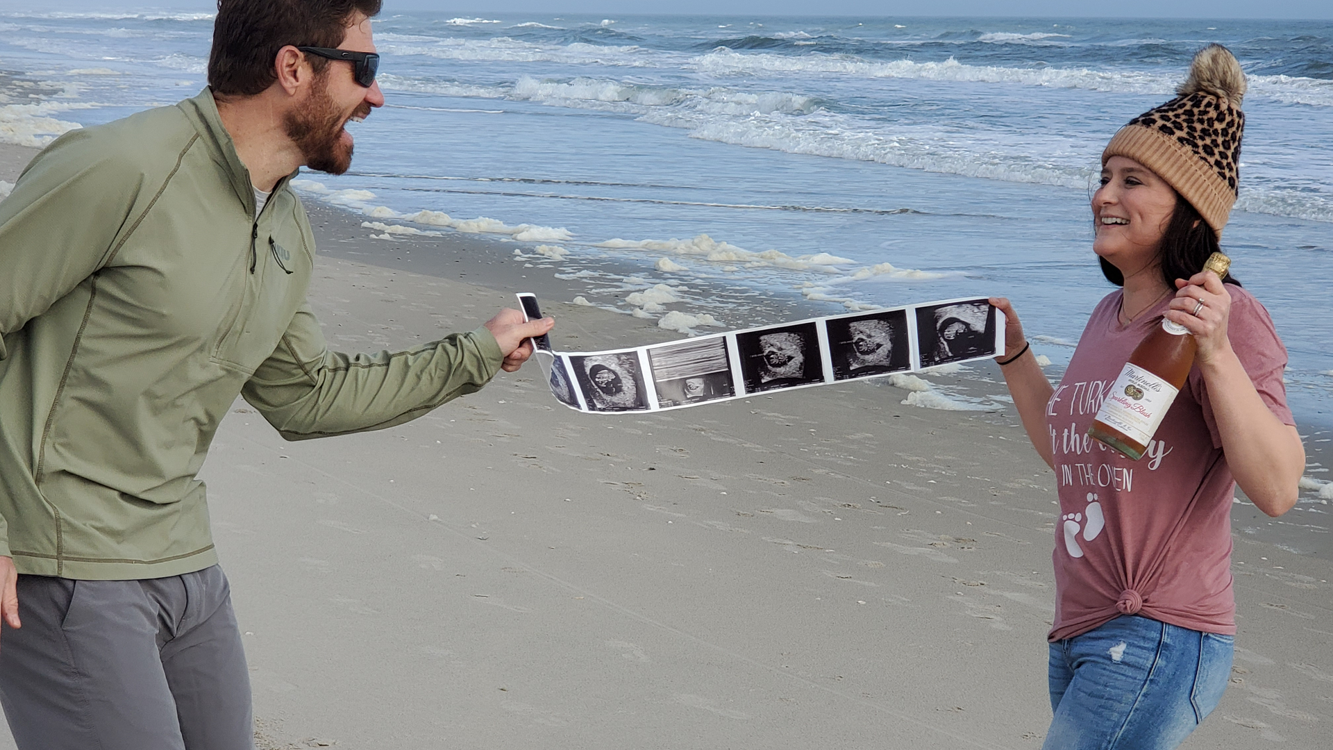 Chelsea and Jake Jovanovich on a sandy beach with a banner of ultrasound images stretched between them