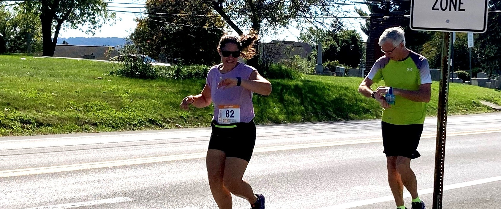 Abbey Oatman and Fred Kohler both look at their fitness trackers while running on a road together on a sunny day