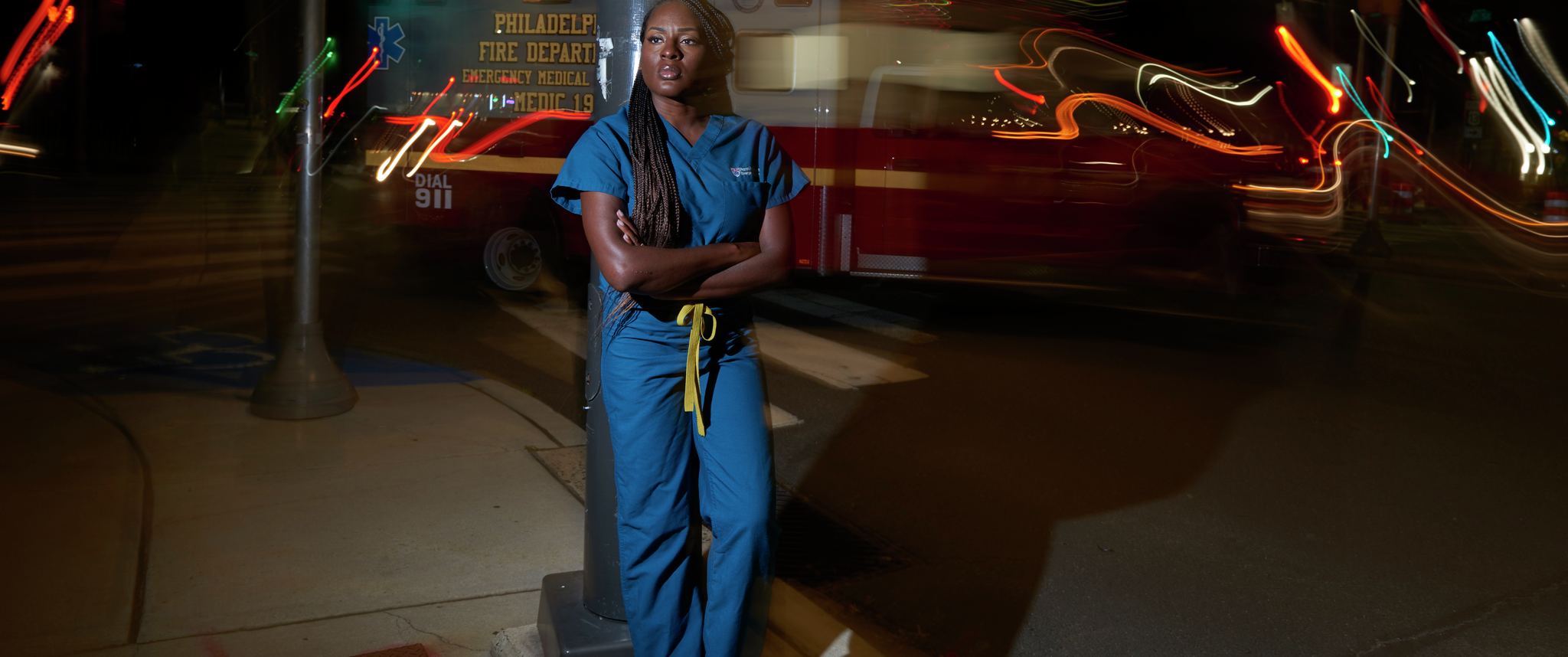 Chidinma Nwakanma, dressed in scrubs, stands outdoors at night looking away, as an ambulance passes behind her