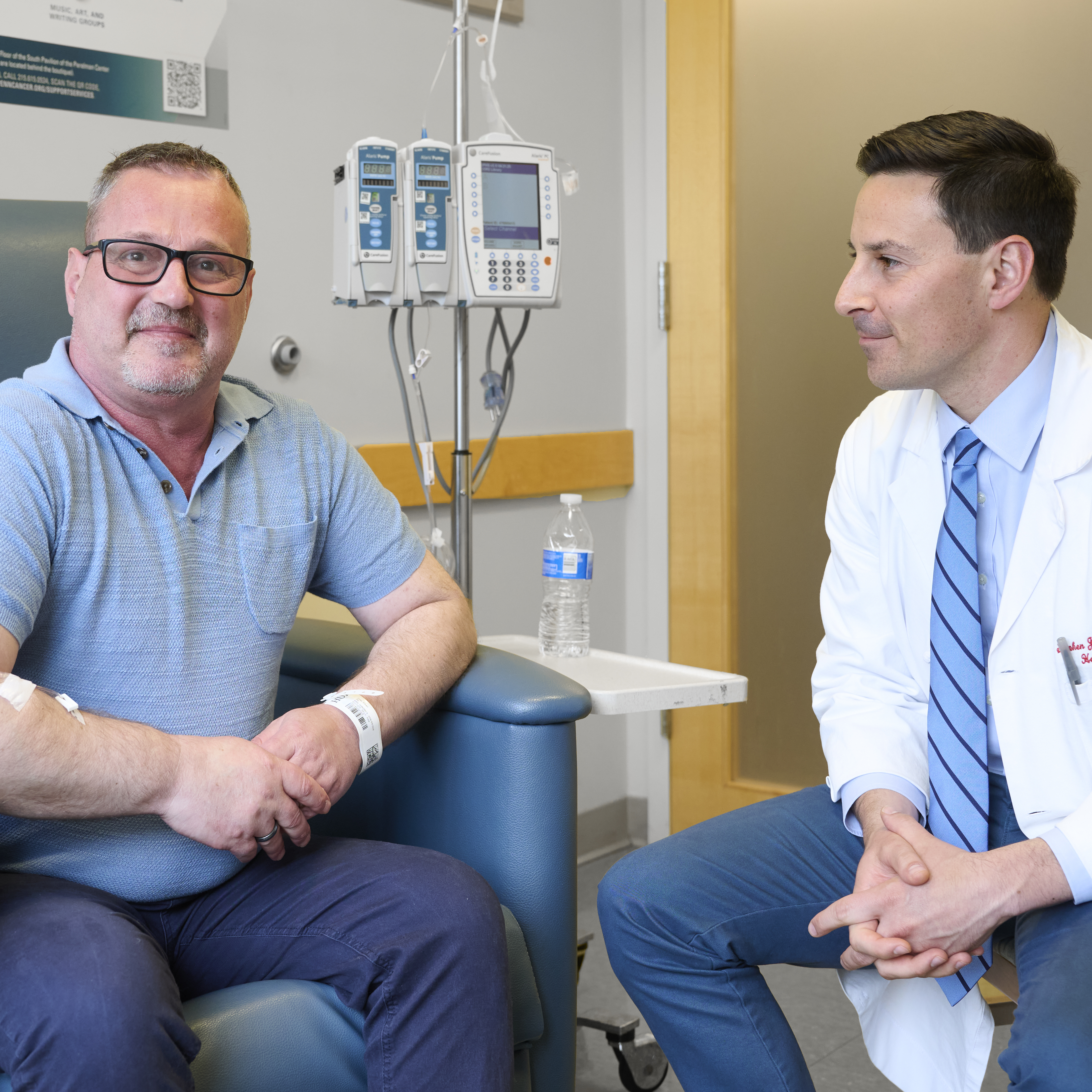 Eduart Cuka, sitting up proud and happy in an infusion chair, with Stephen Bagley, MD, at his side, looking at him 