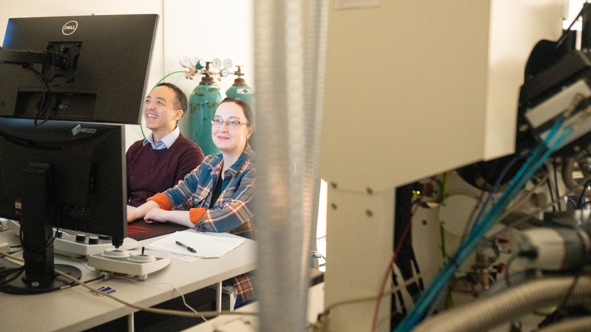 Yi-Wei Chang, PhD, and Katya Tarasovetc, PhD, smile while looking at images on multiple monitors, surrounded by scientific equipment