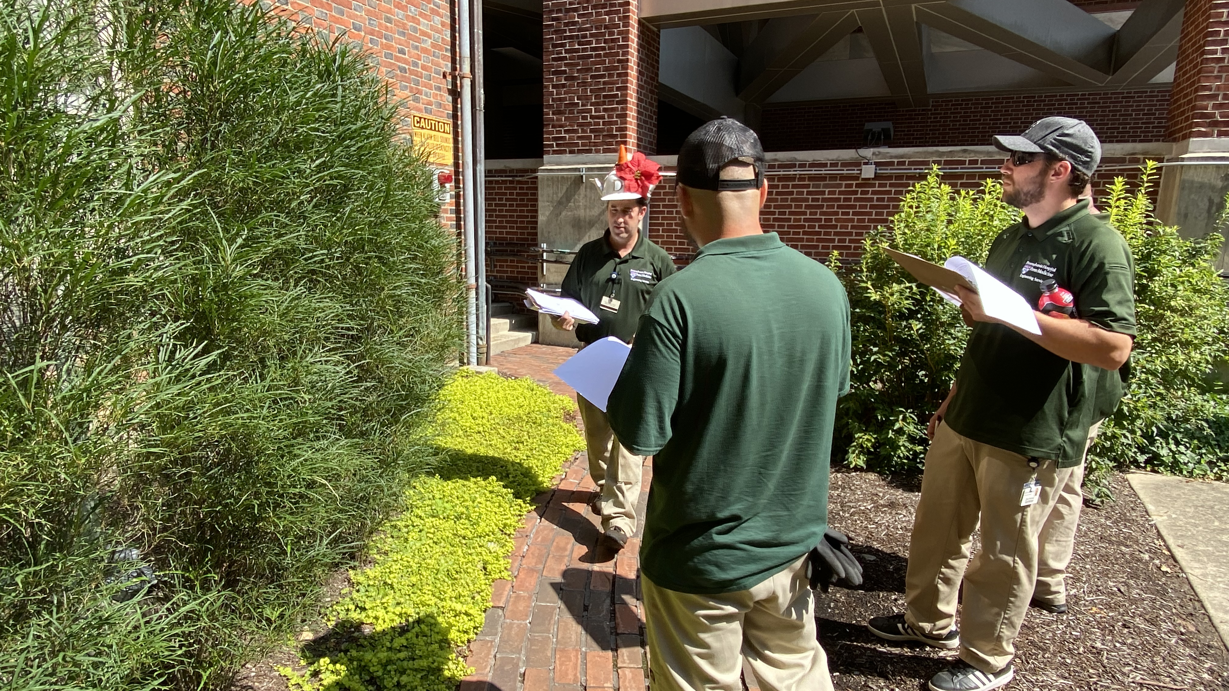 Dan Bangert stands in front of several plants in a courtyard. Nick Sambucetti and Blake Sieminski hold clipboards and examine the plants