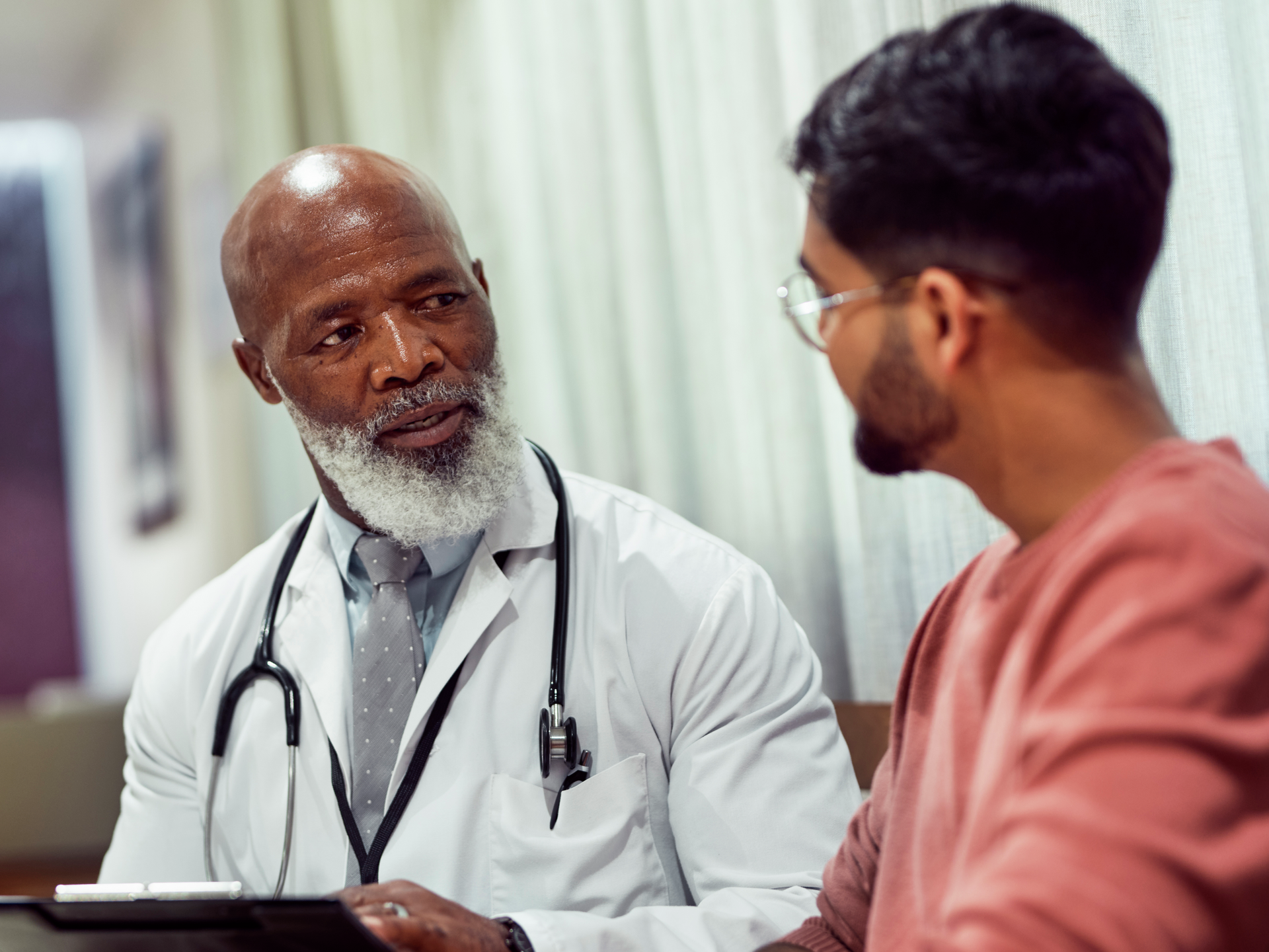 Senior doctor with beard holding clipboard, talking to male patient