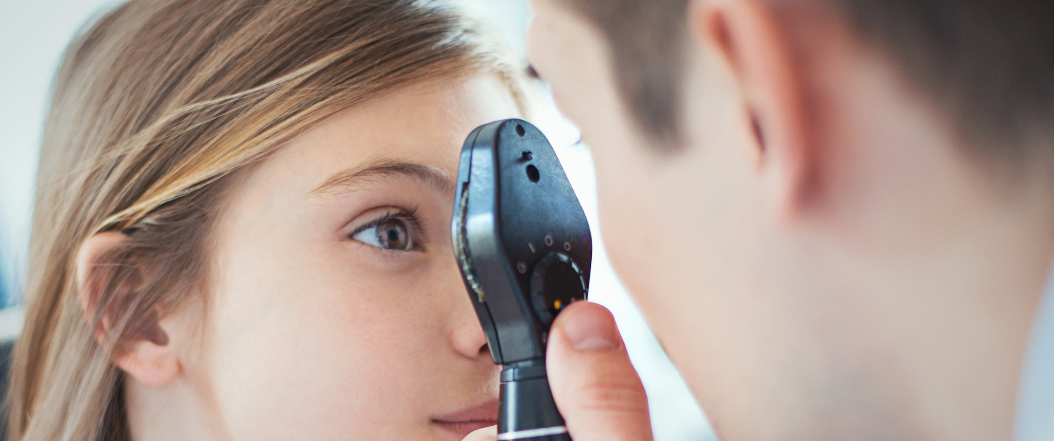 Closeup side view of early 40's unrecognizable optician examining eyesight of his little female patient with a phoropter device. The girls is aged 9, with brown eyes and hair.