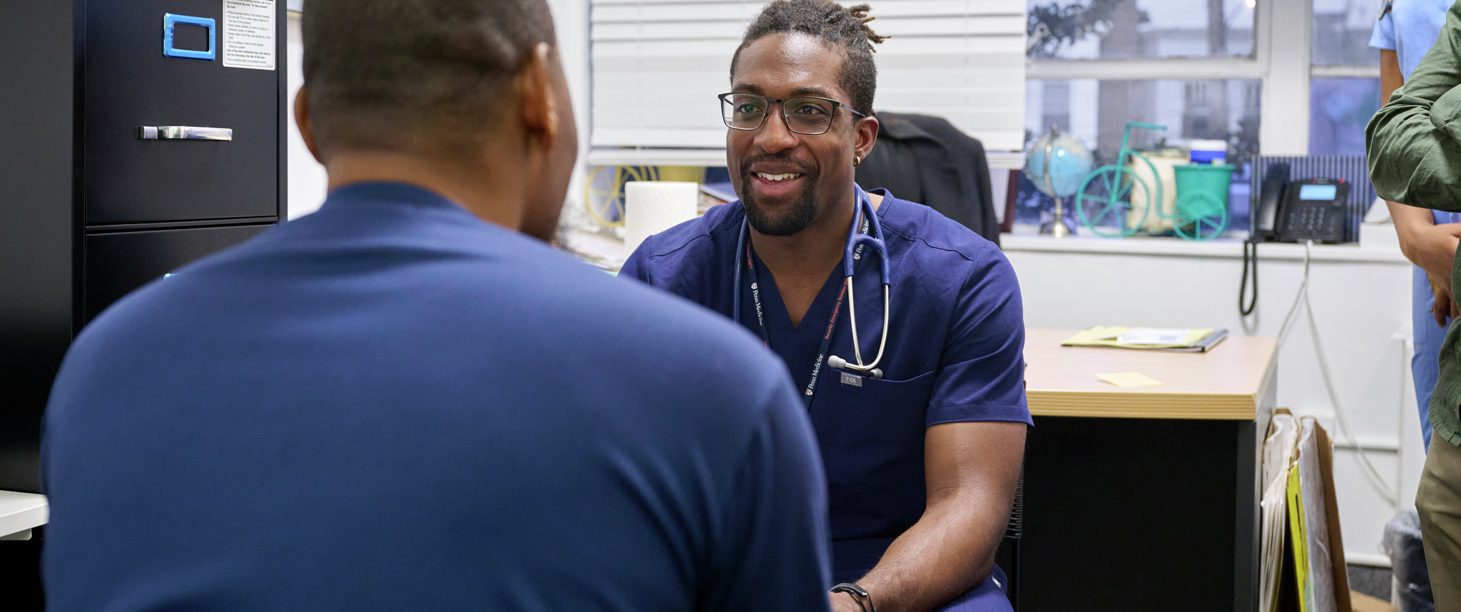 A Penn Medicine staff member in scrubs talks with a patient at the African Health Clinic