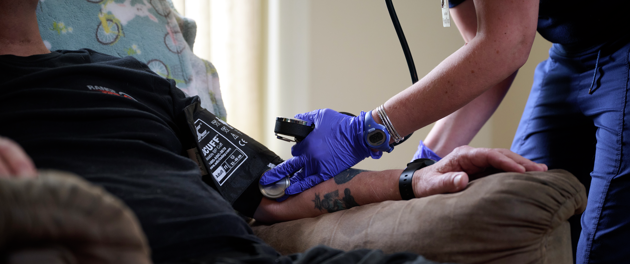 A clinician’s gloved hand presses a stethoscope into the arm of a man seated in a home recliner chair, taking his blood pressure