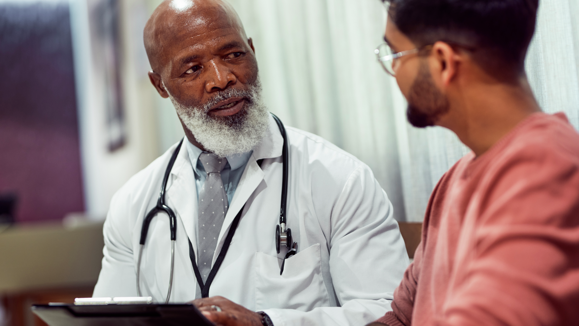 Senior doctor with beard holding clipboard, talking to male patient