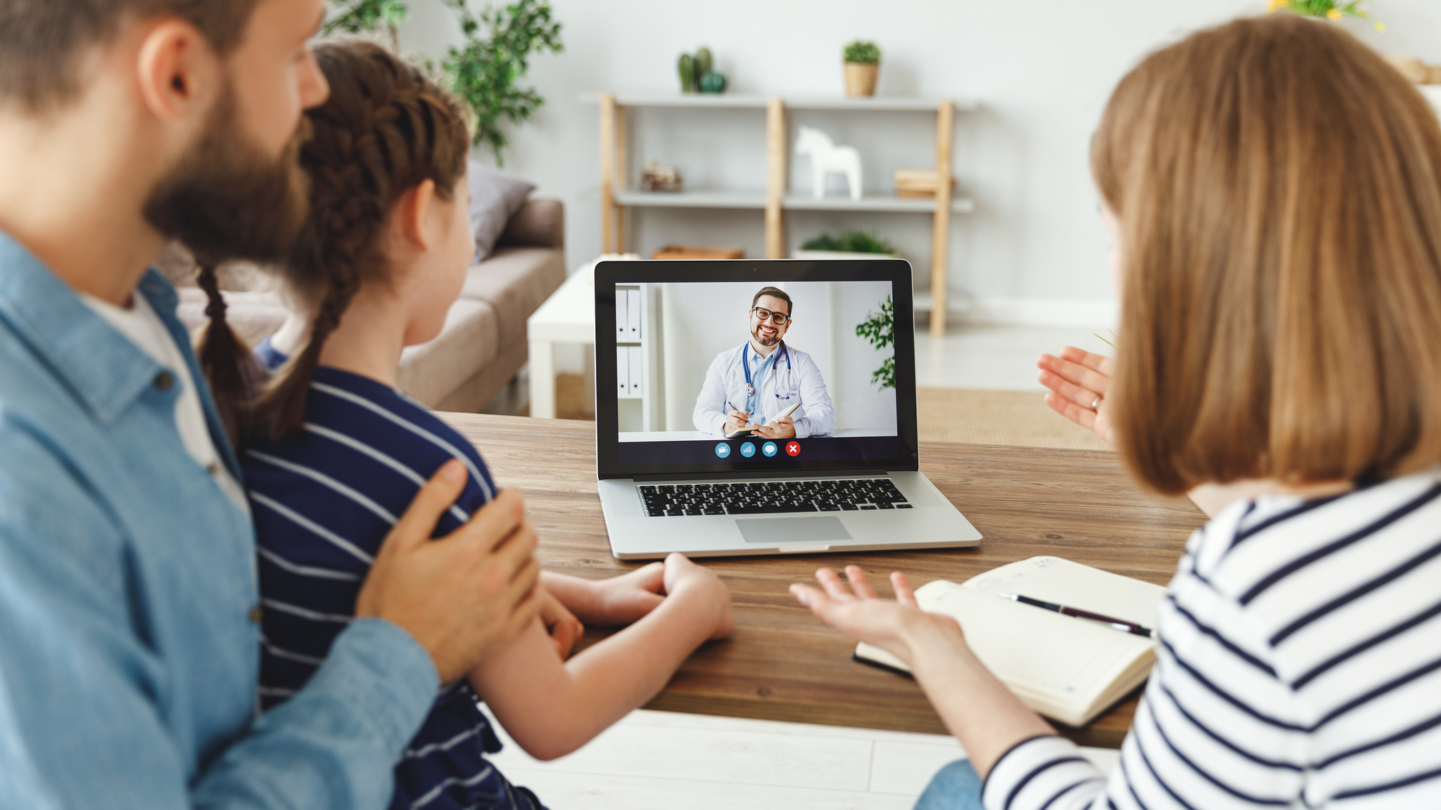 Parents and cheerful daughter sitting at table and speaking with online doctor at home.