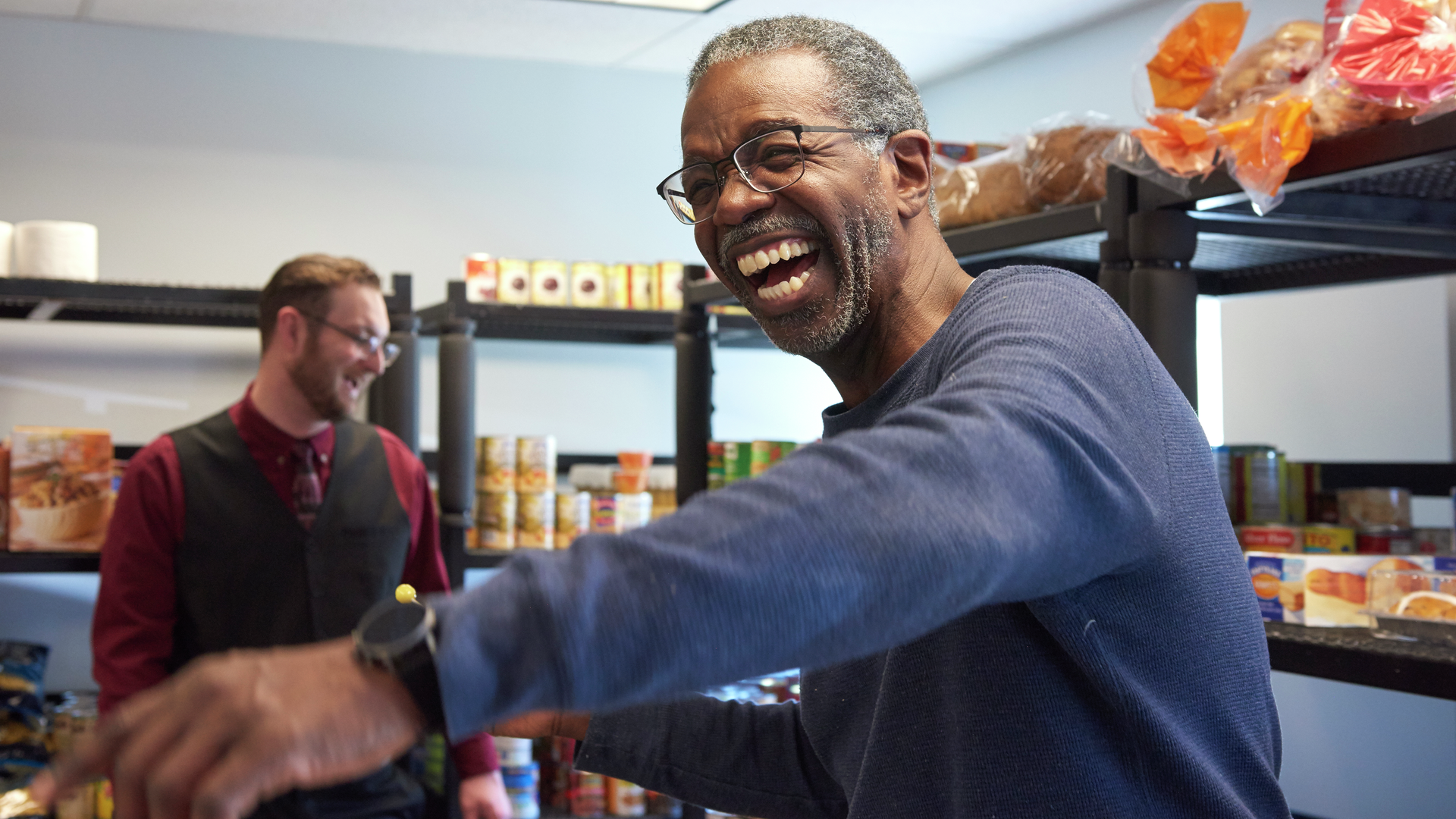 Timmy Nelson smiles big while holding a bag of food in a room lined with shelves; Christopher Moore is in the background