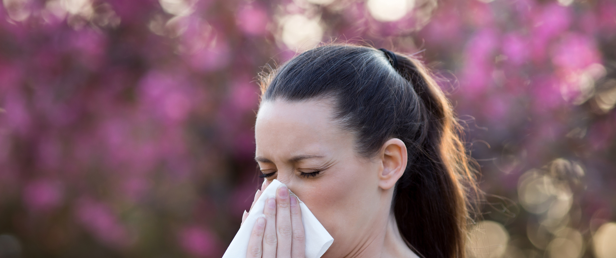 A young woman blows her nose in a tissue with blooming trees in the background
