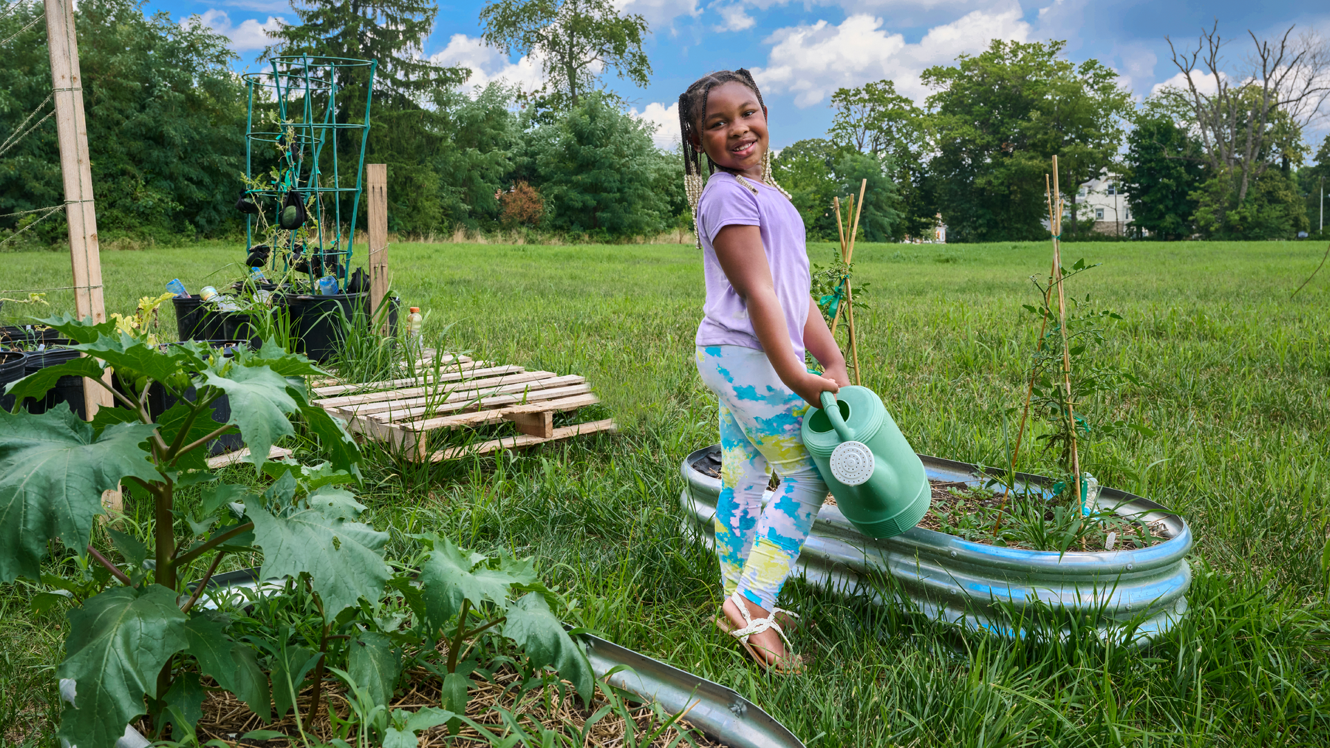 A little girl with braids smiles and poses holding a watering can