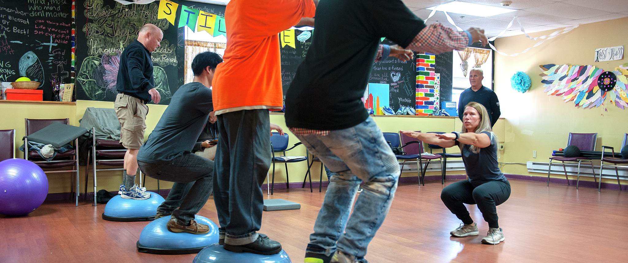 Several men of varied ages and ethnicities practice squatting on uneven rubber round surfaces, guided by a female instructor