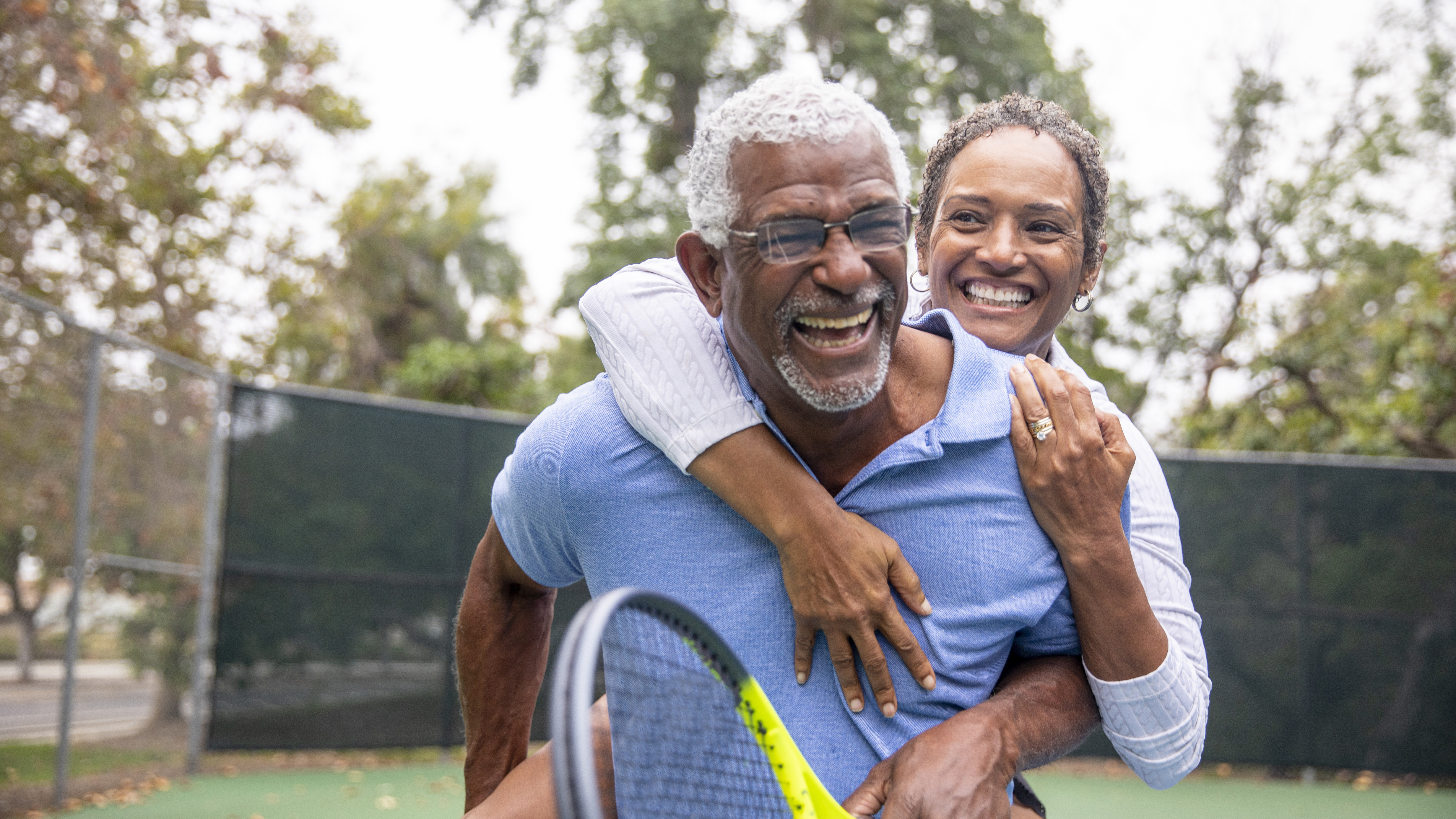 A senior black couple piggyback together on the tennis court