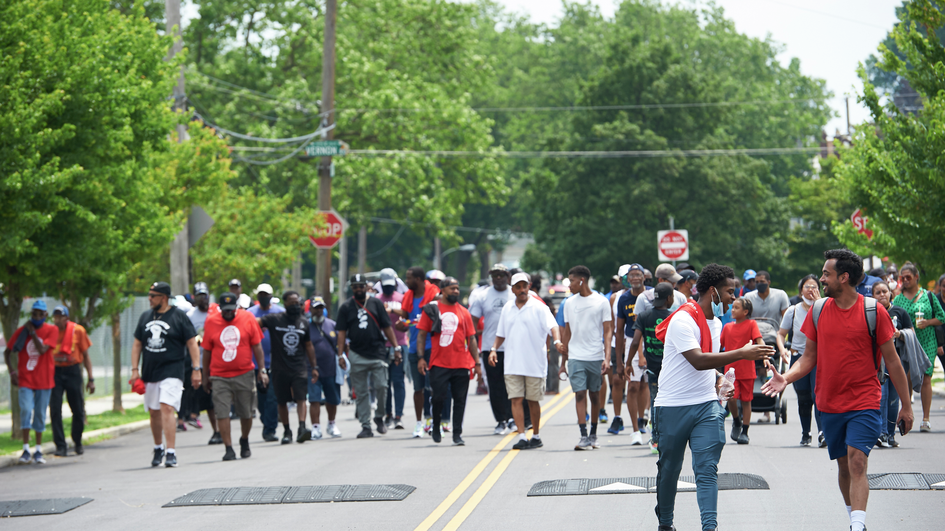 Black men of varied ages wearing red, black, and white T-shirts walk down a road as part of a community walk event