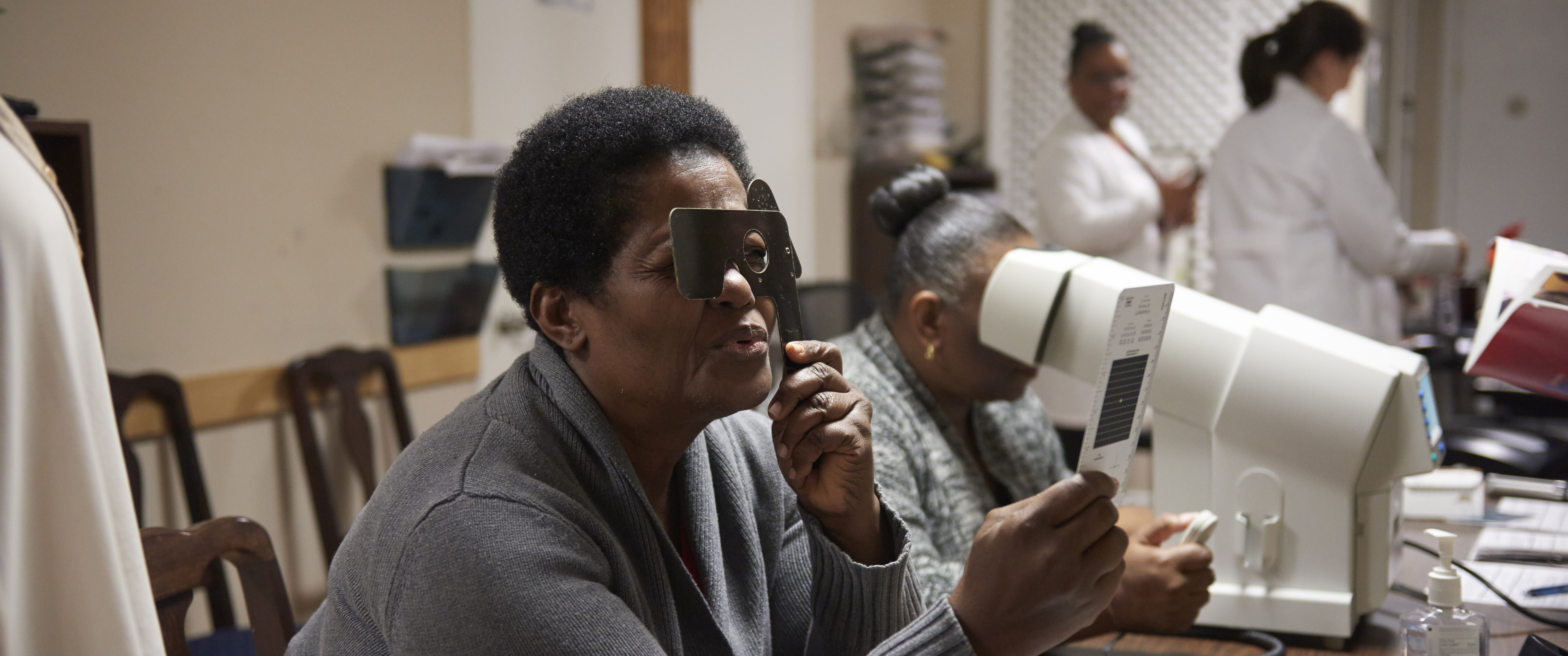 A Black woman holds up a card and a device blocking one eye, for a vision exam, in a room with doctors and patients