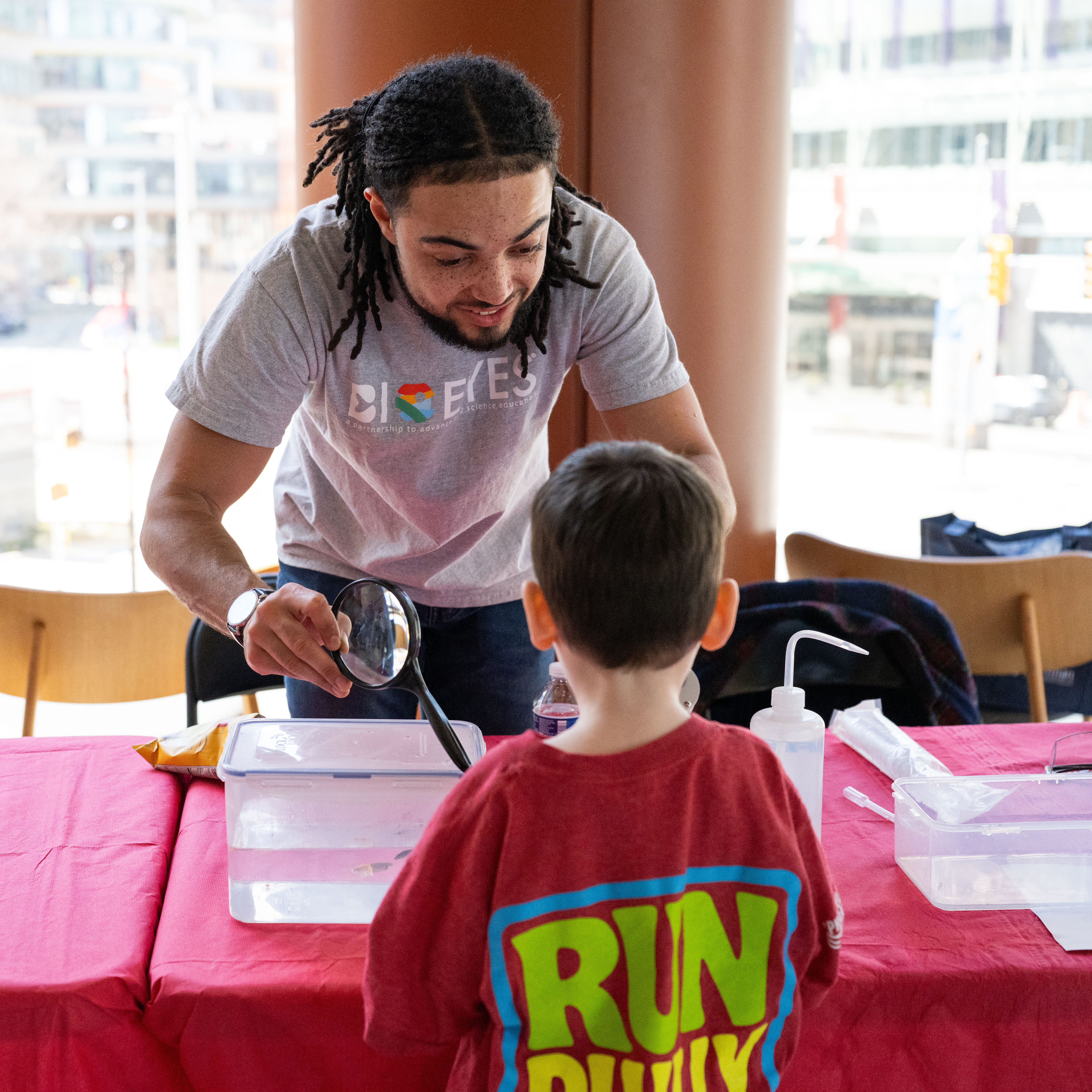 Joshua Kouassi handing a magnifying glass to a child to look at a zebrafish tank at the BioEYES table at a Penn Medicine event