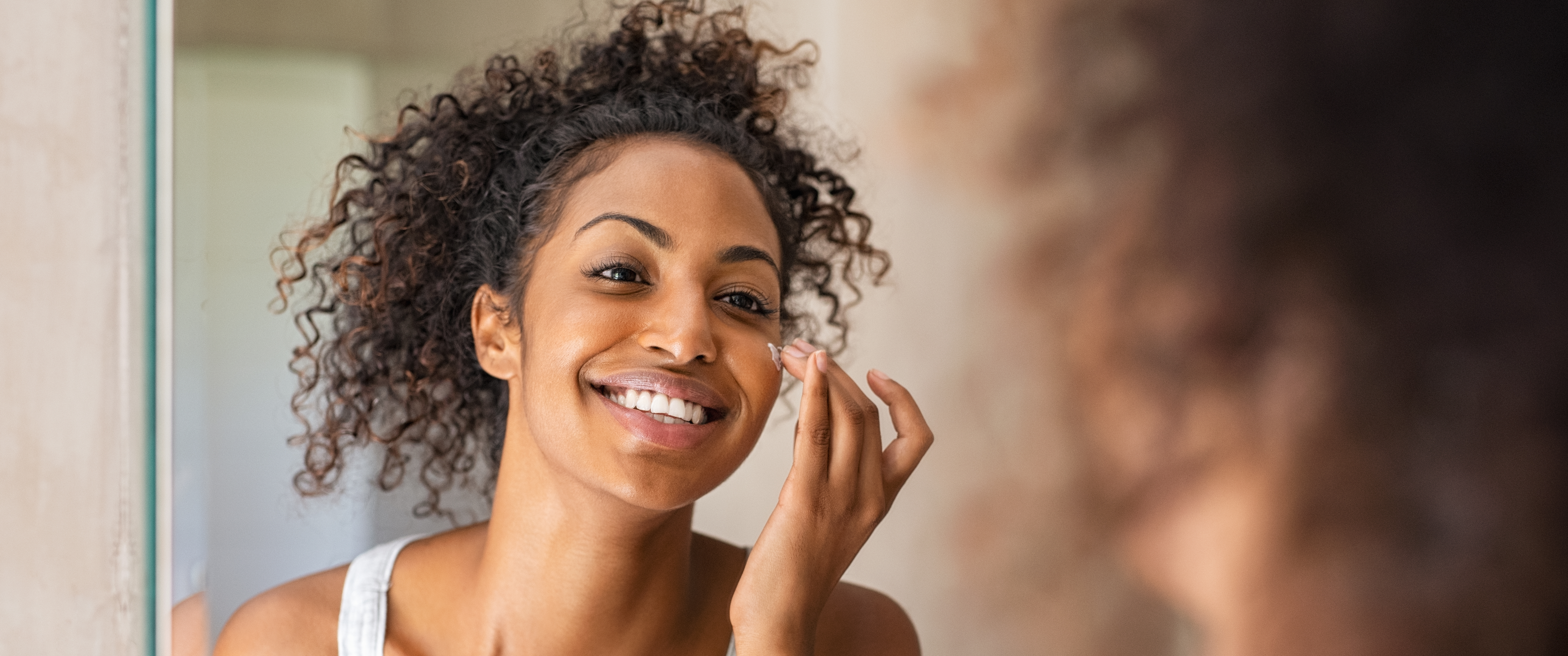 Young african woman applying moisturizer on her face while standing in front of the mirror