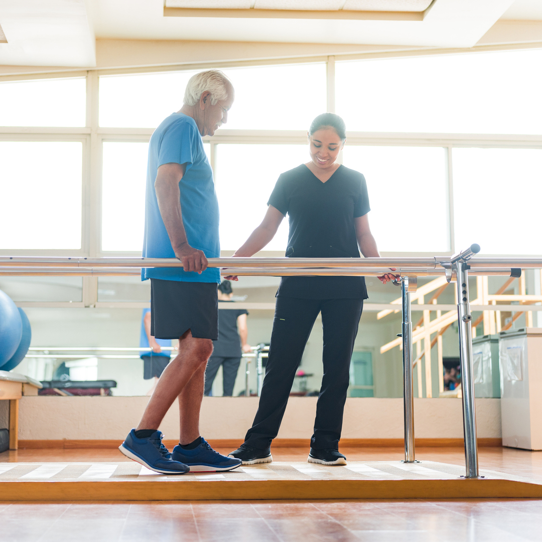 A senior man giving small steps in the parallel bars and physical therapist standing next to him.