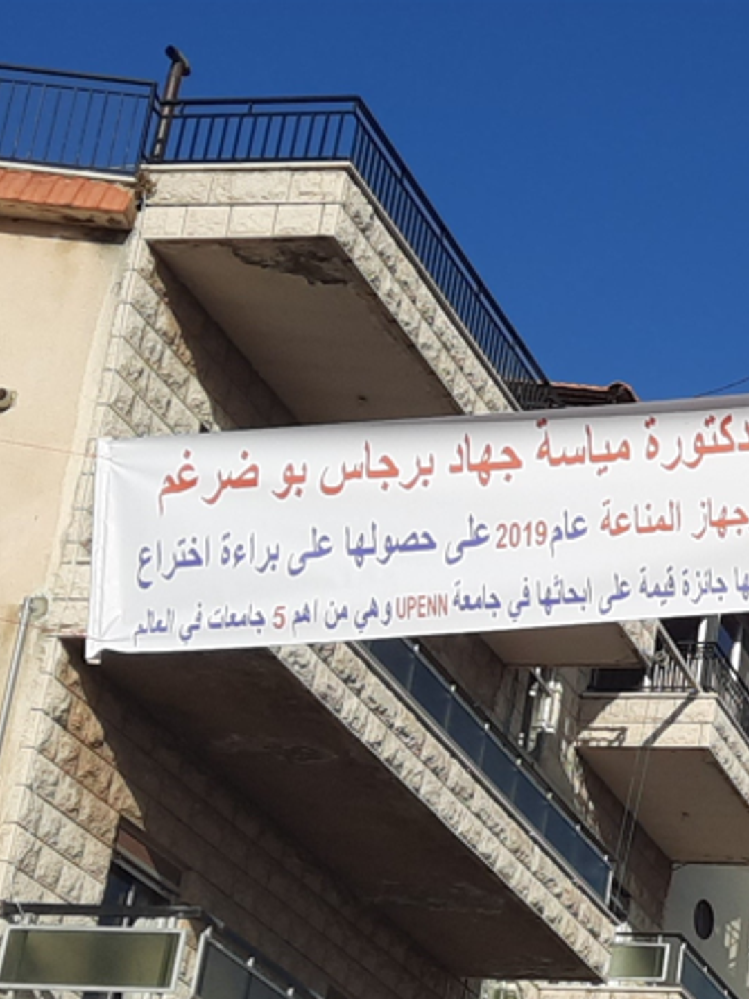 A banner hanging between buildings with Arabic text that congratulates Mayassa Burjas Bou Dargham for her research achievements.