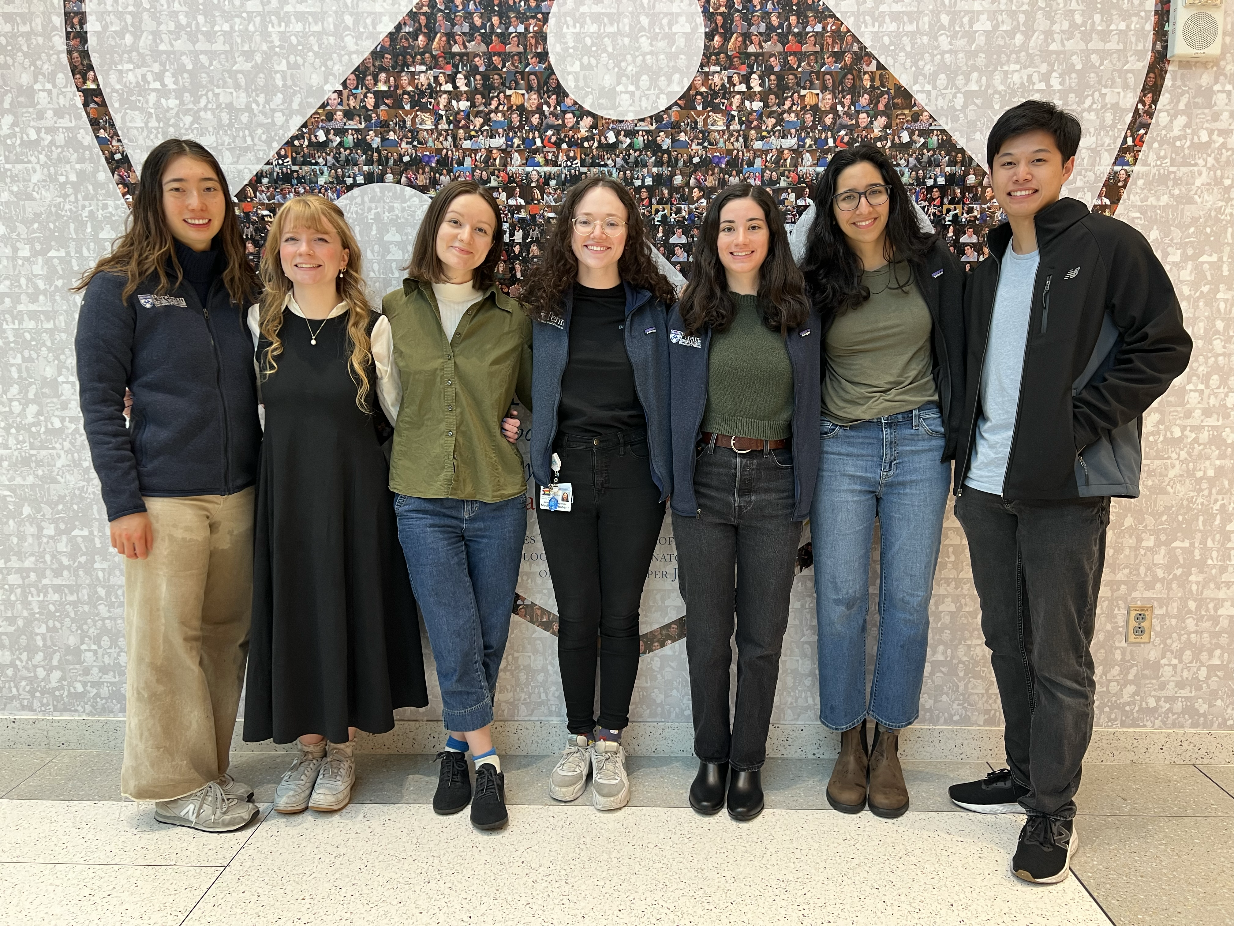 A group photo of medical students Catherine Shen, Elizabeth Whidden, Phoebe Cunningham, Jess Campanile, Sarah Wornow, Isha Thapar, and Jonathan Szeto standing in front of a photo collage in the shape of the Penn shield.