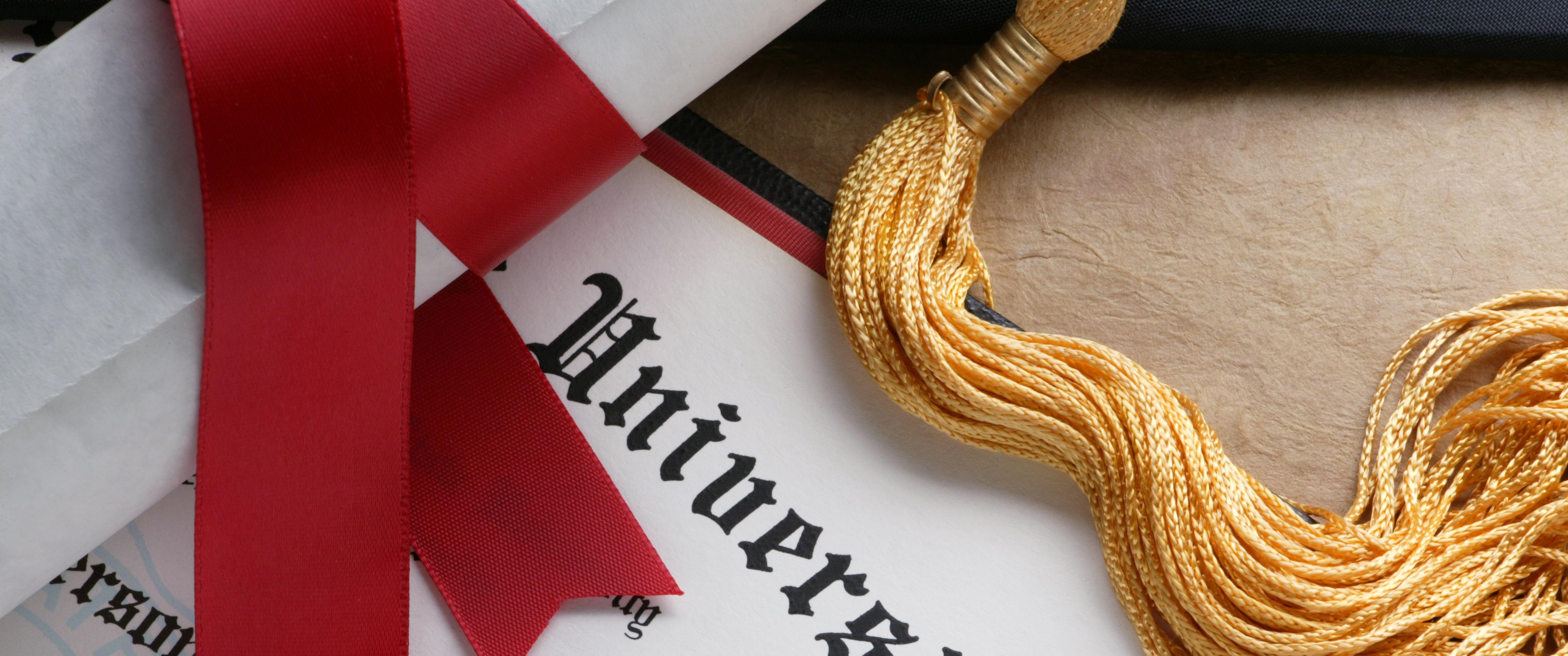 A red ribbon wrapped around a rolled up diploma sits on top of a another college diploma. A gold tassel from a graduation cap is also draped across the diploma.