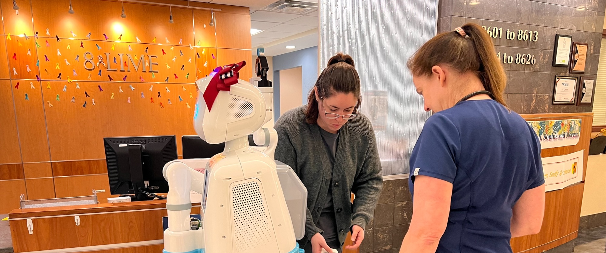 Two nurses interact with a human-sized white robot wearing a red bow atop her head
