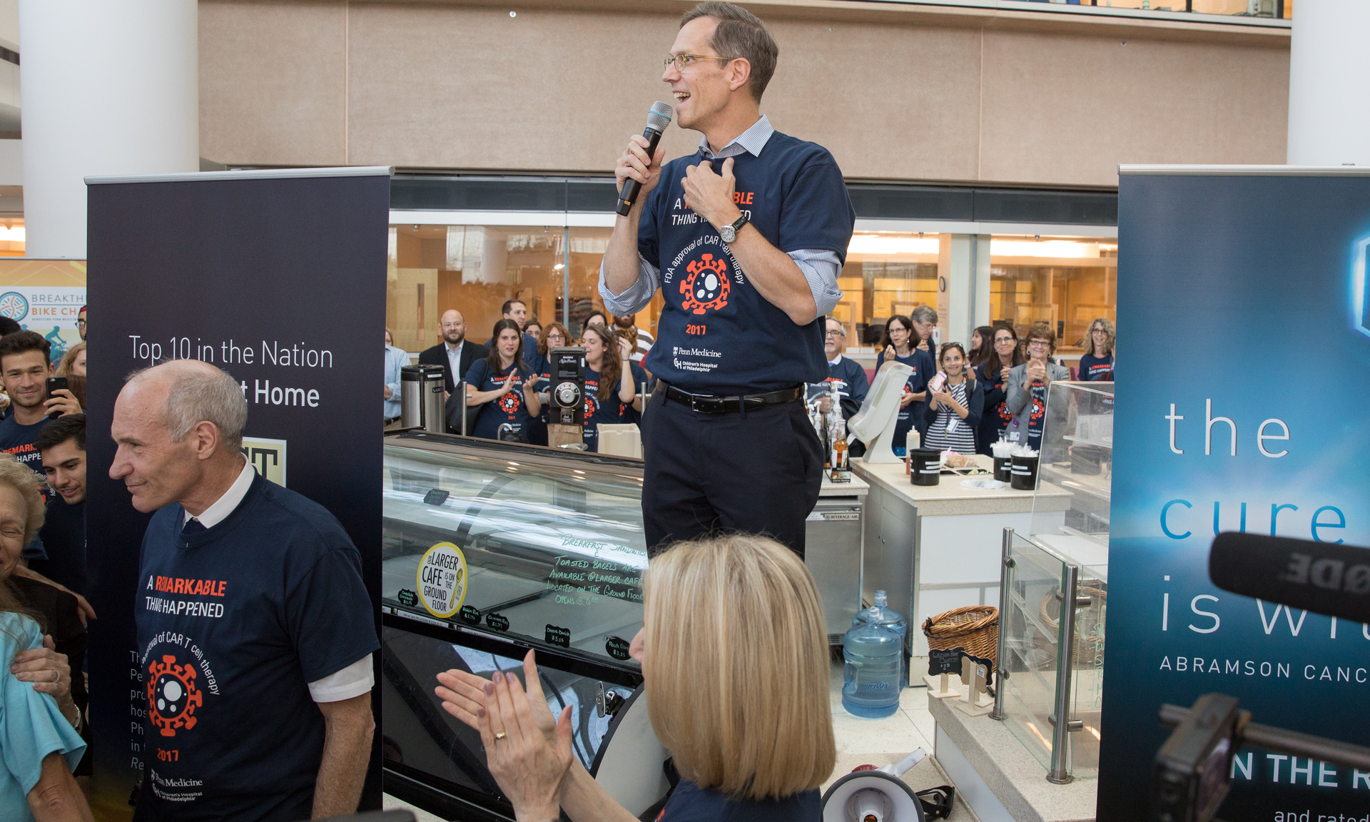Robert Vonderheide stands atop a coffee stand in the Perelman Center, holding a microphone and speaking to a crowd with Carl June in the foreground