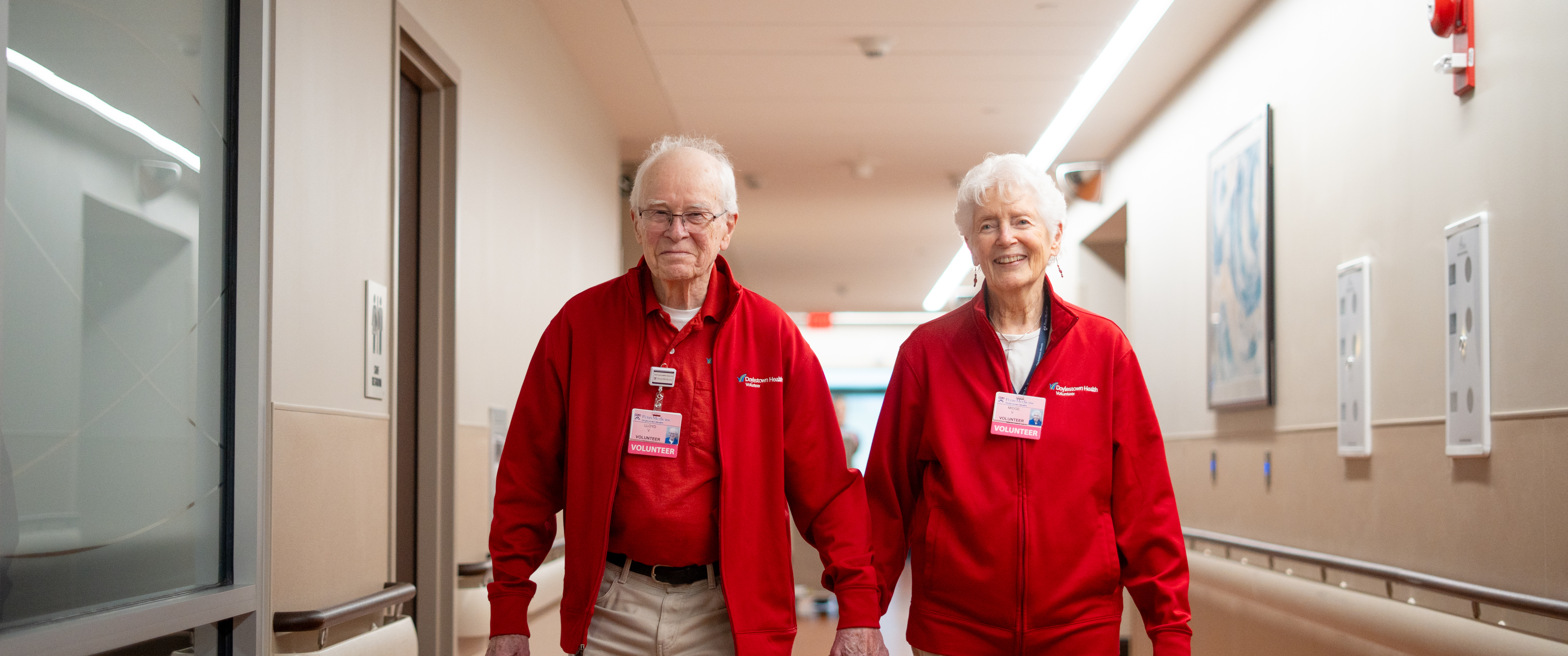 Lloyd and Midge smile at the camera as they walk hand-in-hand through a Doylestown Hospital hallway. They are wearing matching tan khaki pants and matching red sweatshirts