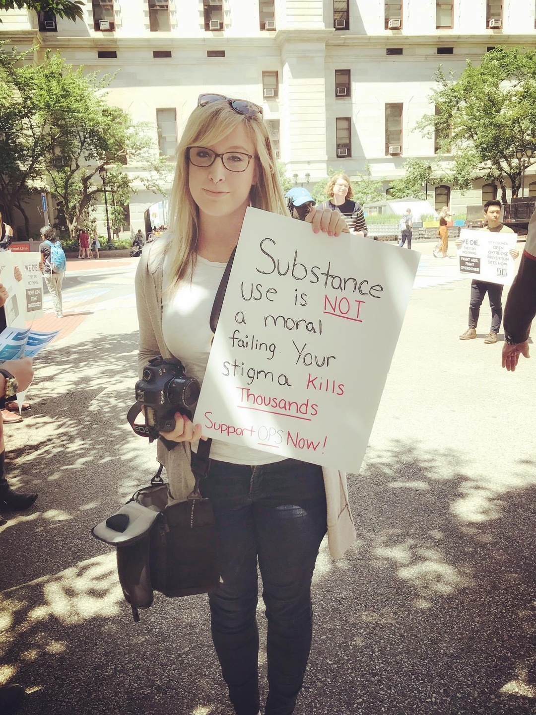 Brittany Salerno stands holding a camera and a sign that says, "Substance use is not a moral failing. Your stigma kills thousands."