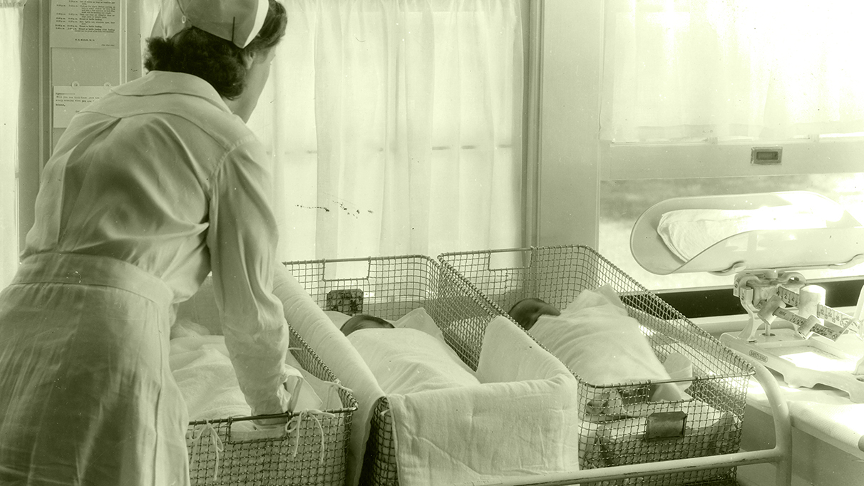 Old photograph of a female nurse tending to three babies in wire baskets, next to a scale.