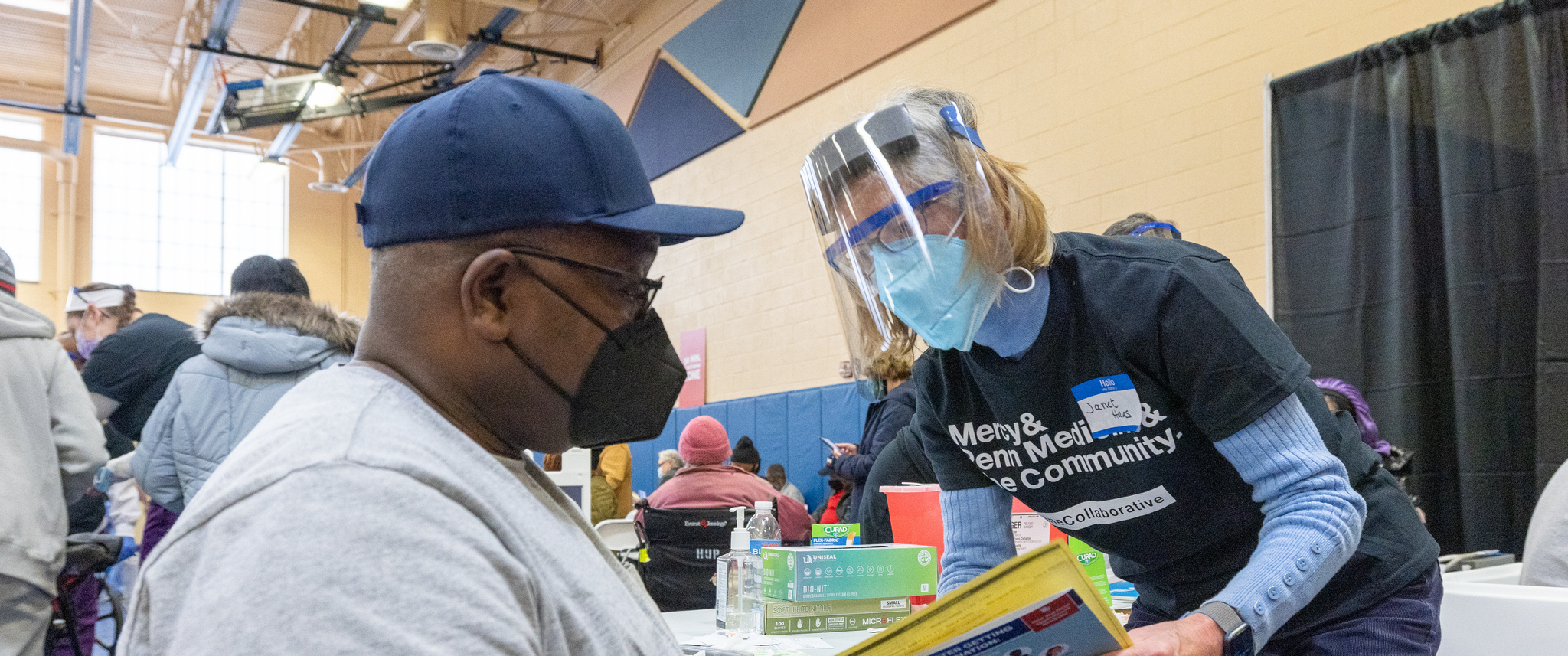 A woman wearing a navy T-shirt, mask, and face shield leans down to talk with a seated masked man
