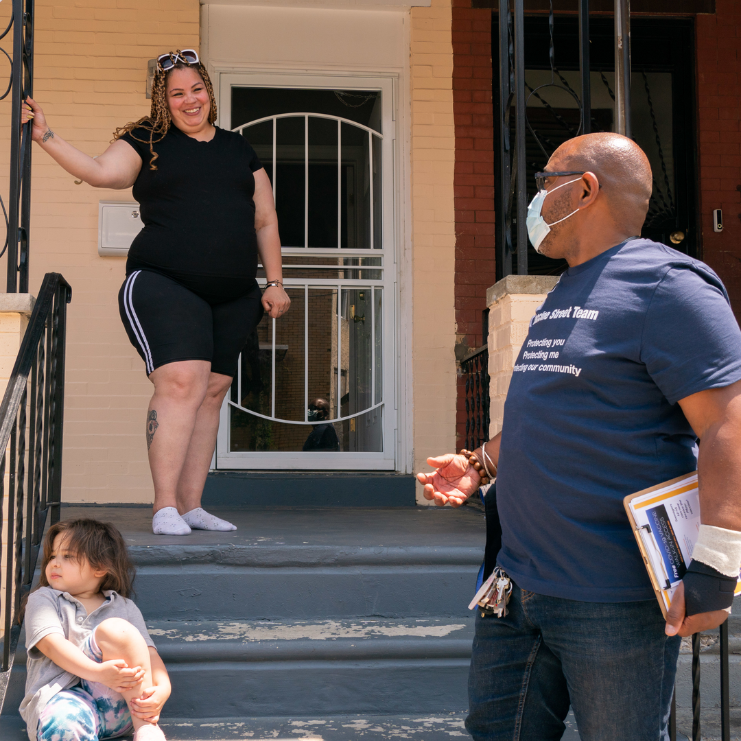 A young woman smiles looking down her front porch steps at Yuhnis Syndor, while a small child plays