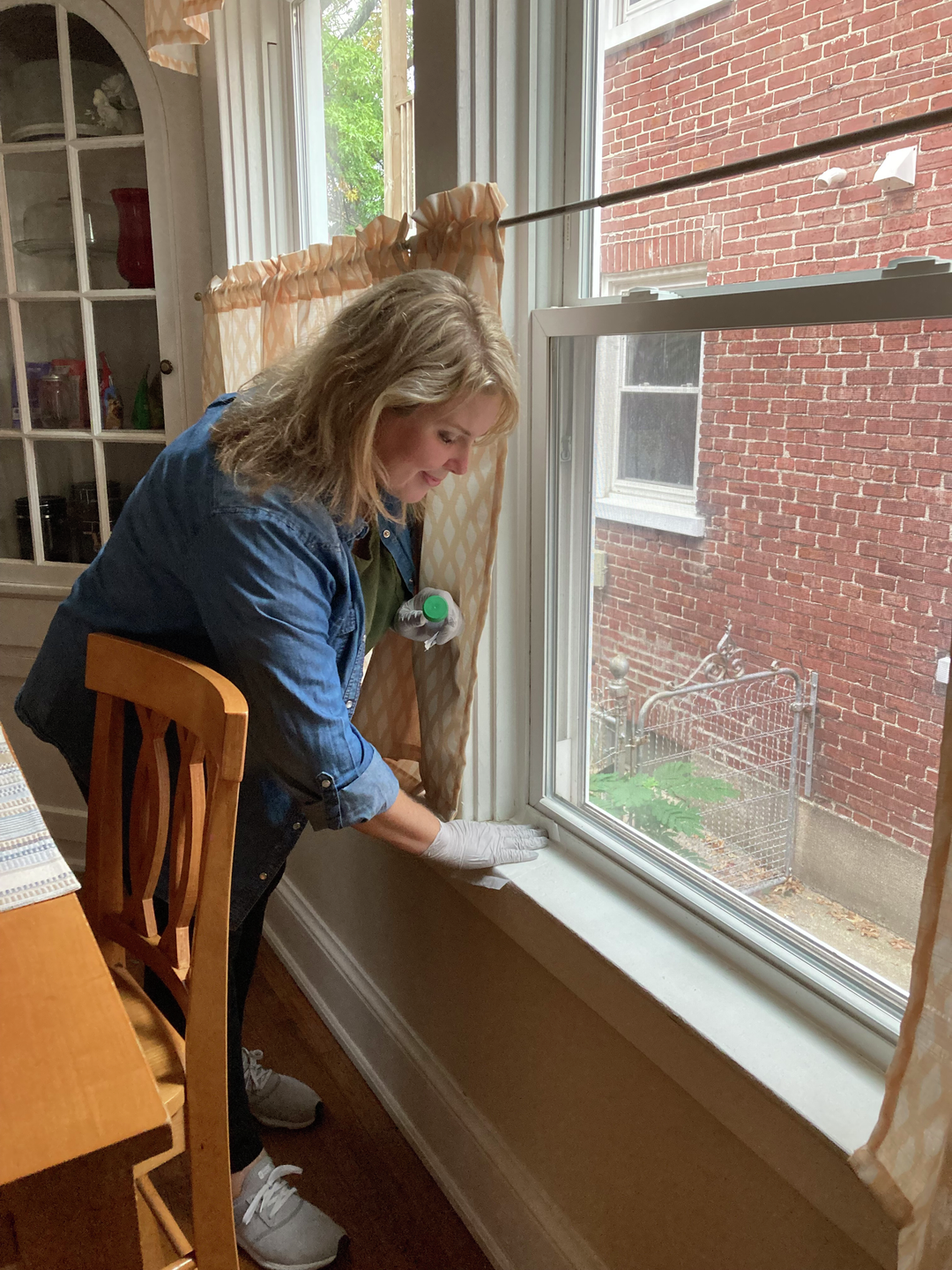 A woman wipes down a window sill, with the brick exterior visible through the window.