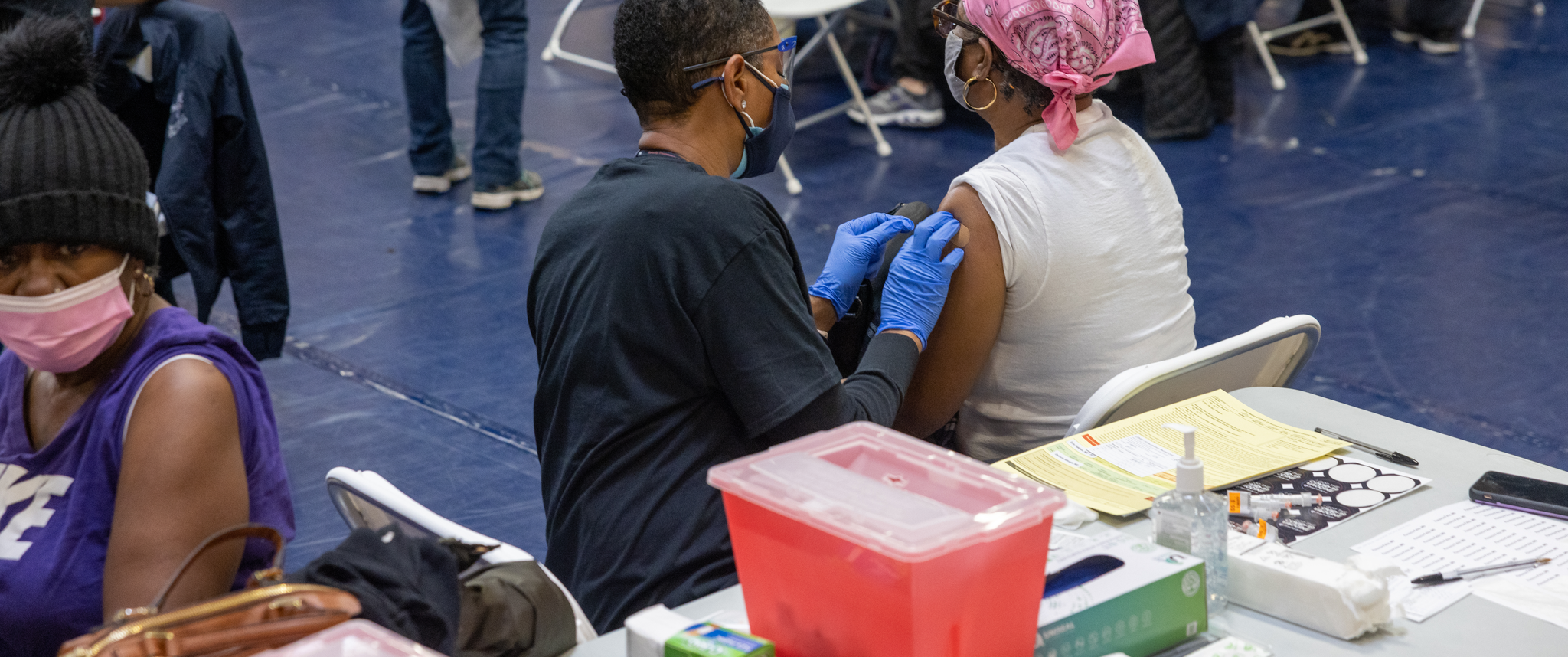In a crowded gymnasium filled with folding tables and chairs, a Black woman wearing blue gloves puts a bandage on the upper arm of a Black woman who just received a COVID vaccine.