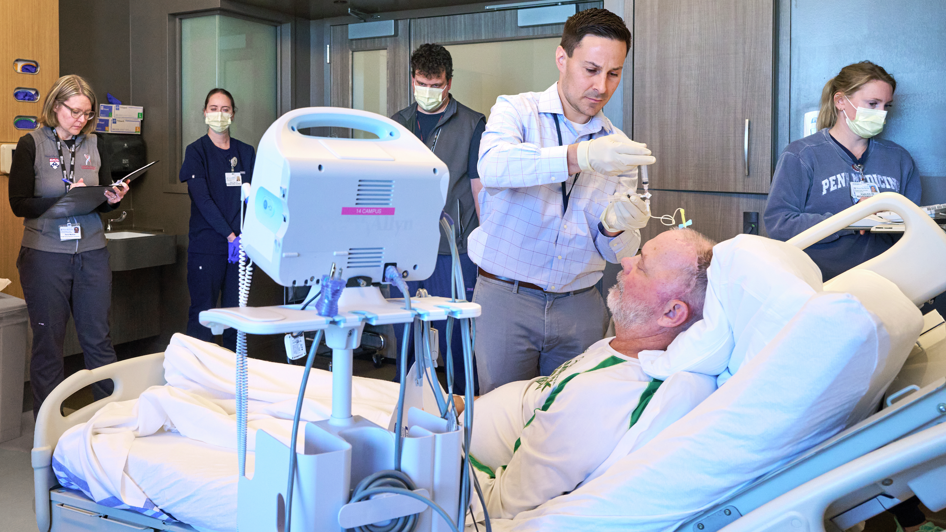Stephen Bagley, MD, injecting CAR T cells into a male patient in a hospital bed while clinical research staff stand nearby observing and taking notes