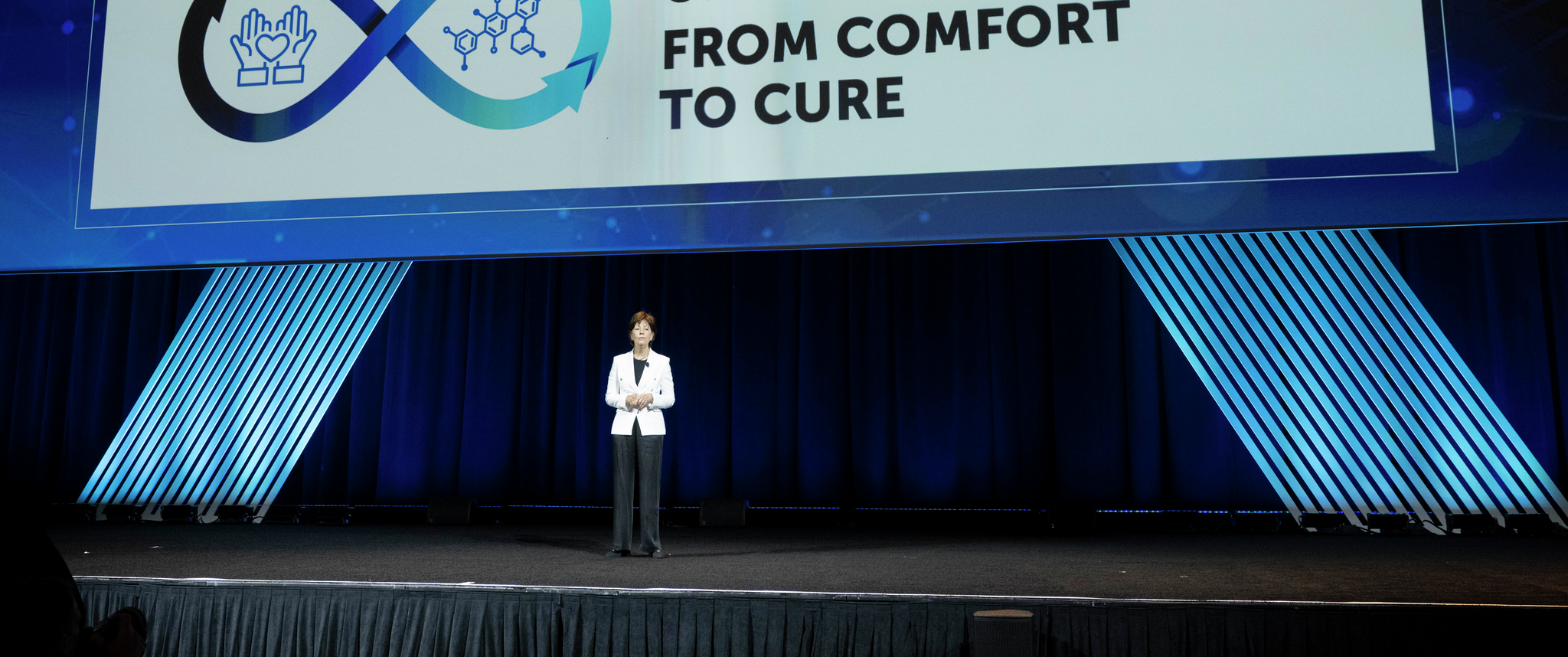 Lynn Schuchter stands on a stage, a large screen behind her reading: ASCO 2024, The Art and Science of Cancer Care: From Comfort to Cure