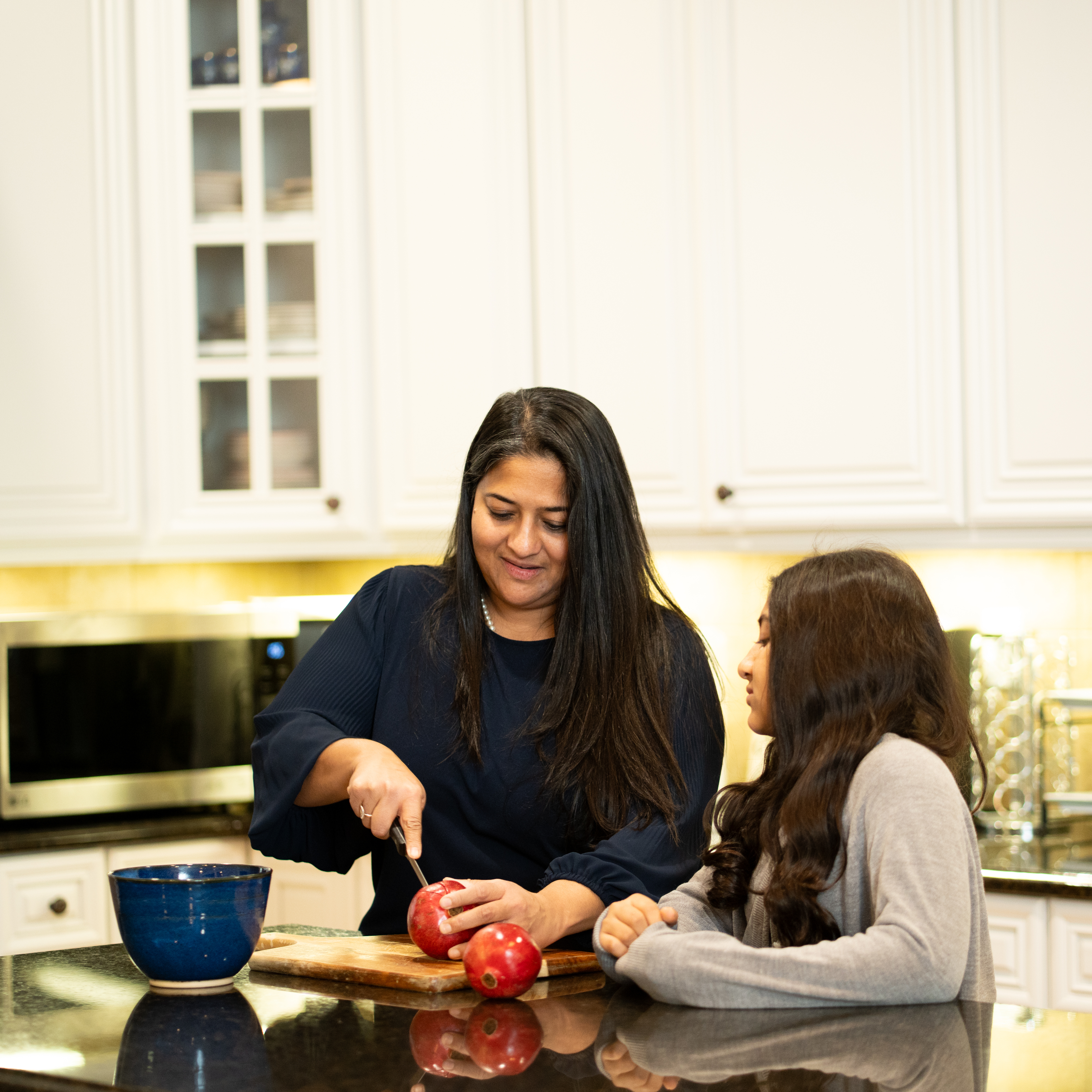 Aileen John and her daughter standing in their kitchen, cutting a pomegranate on the counter to prepare for a meal