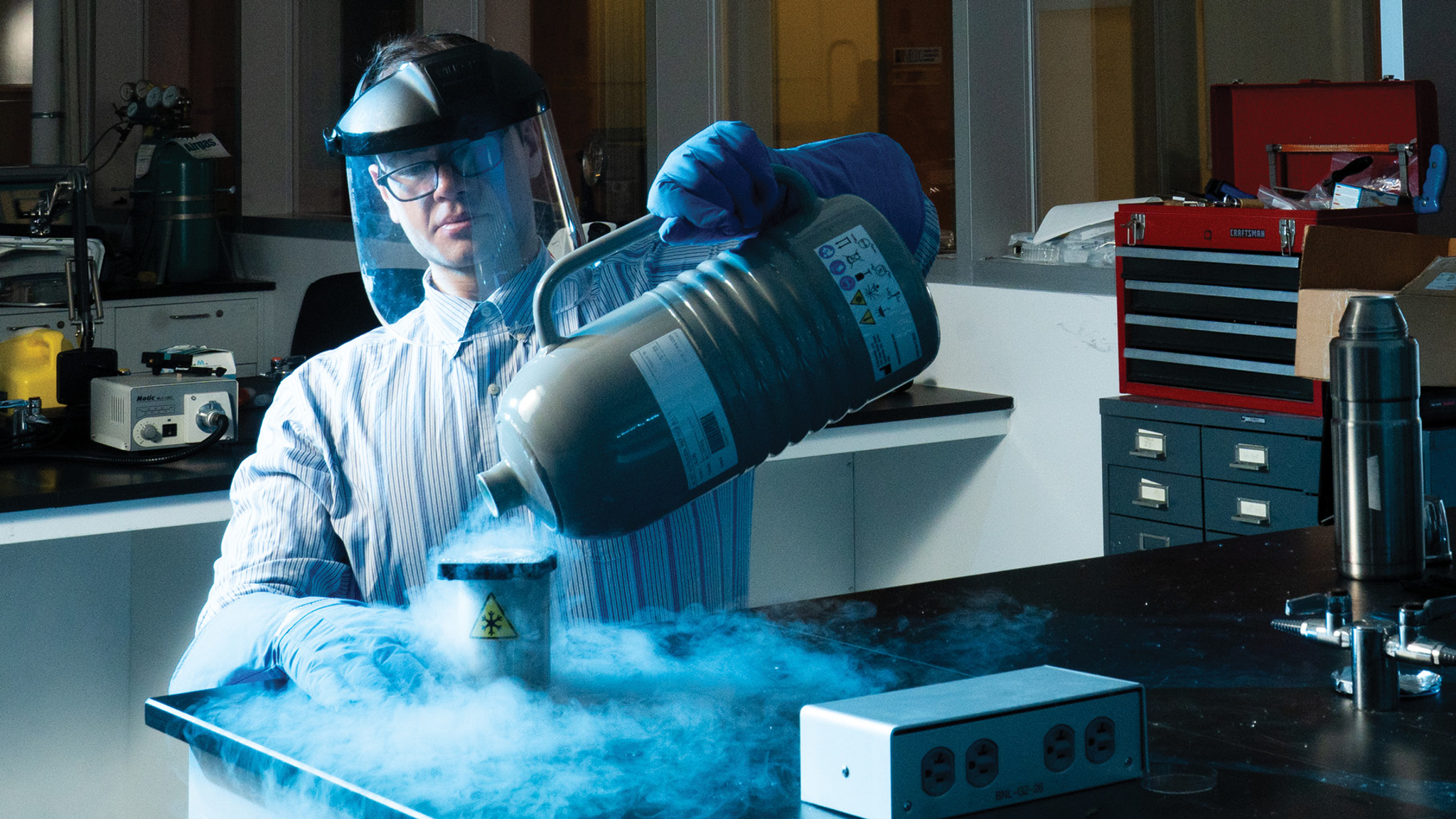 Nicholas Palmer, wearing a face shield and thick gloves, pours cold liquid nitrogen from a jug into a vessel with a snowflake symbol in a coolly lit lab
