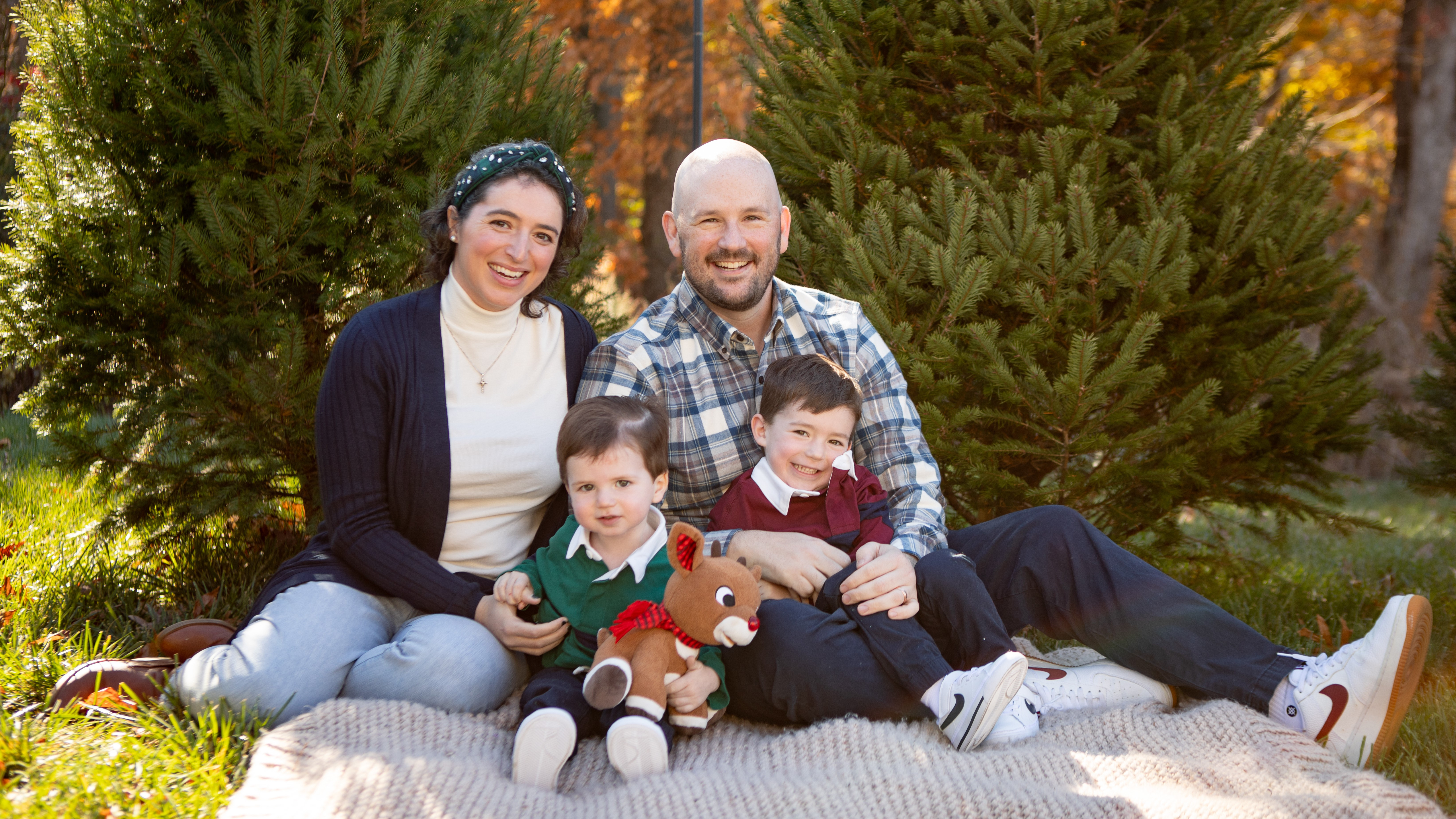 Family of four sitting on a blanket outdoors in front of evergreen trees, smiling. 