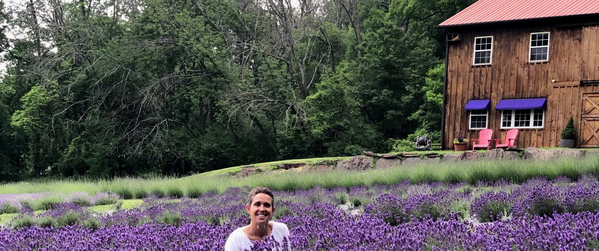 Patti Lyons kneels in a large field of lavender blossoms that reach her neck, with a wooded area and cozy cabin behind her.