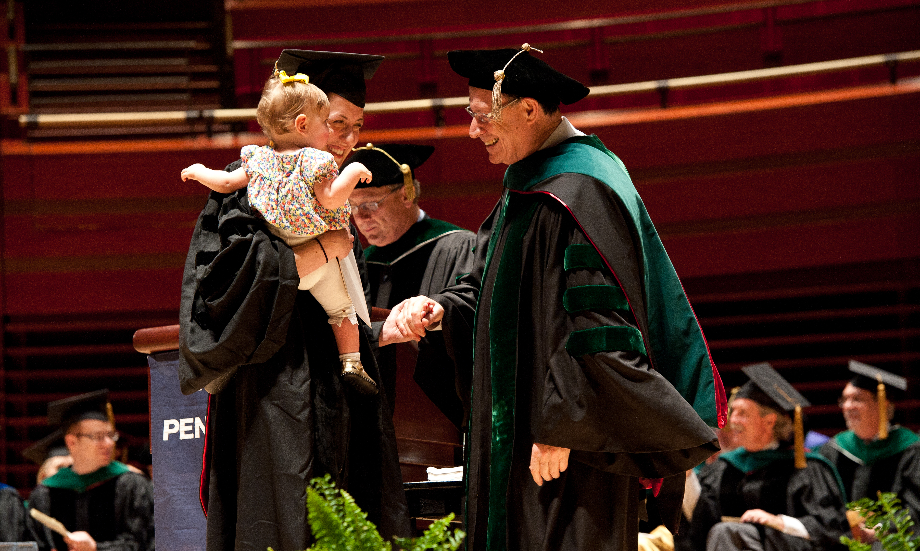 Arthur Rubenstein warmly shakes the hand of a female medical school graduate, who holds a baby, at the commencement ceremony
