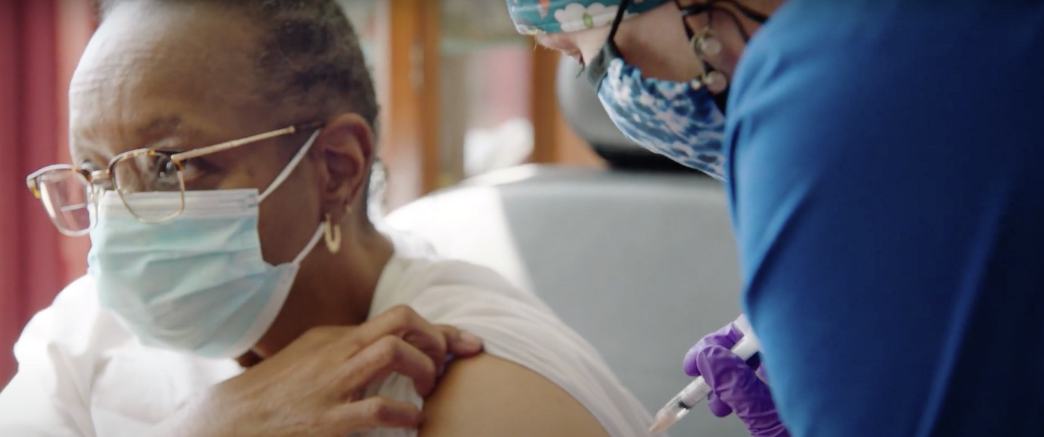 An older Black woman wearing a face mask lifts her sleeve for a nurse to give her a vaccine shot