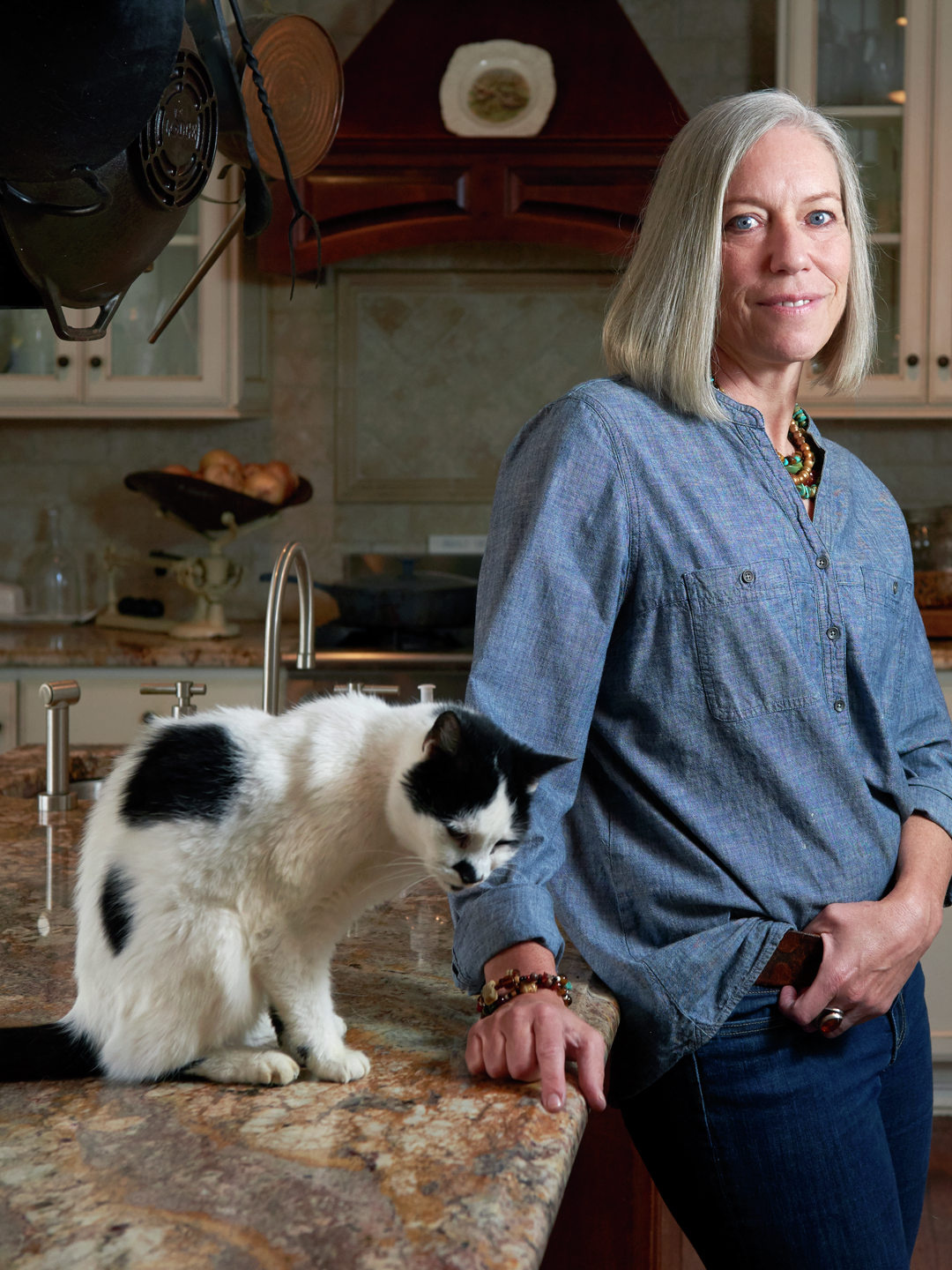 Liz Cleary stands with a slight smile in her kitchen with a cat sitting beside her on the countertop
