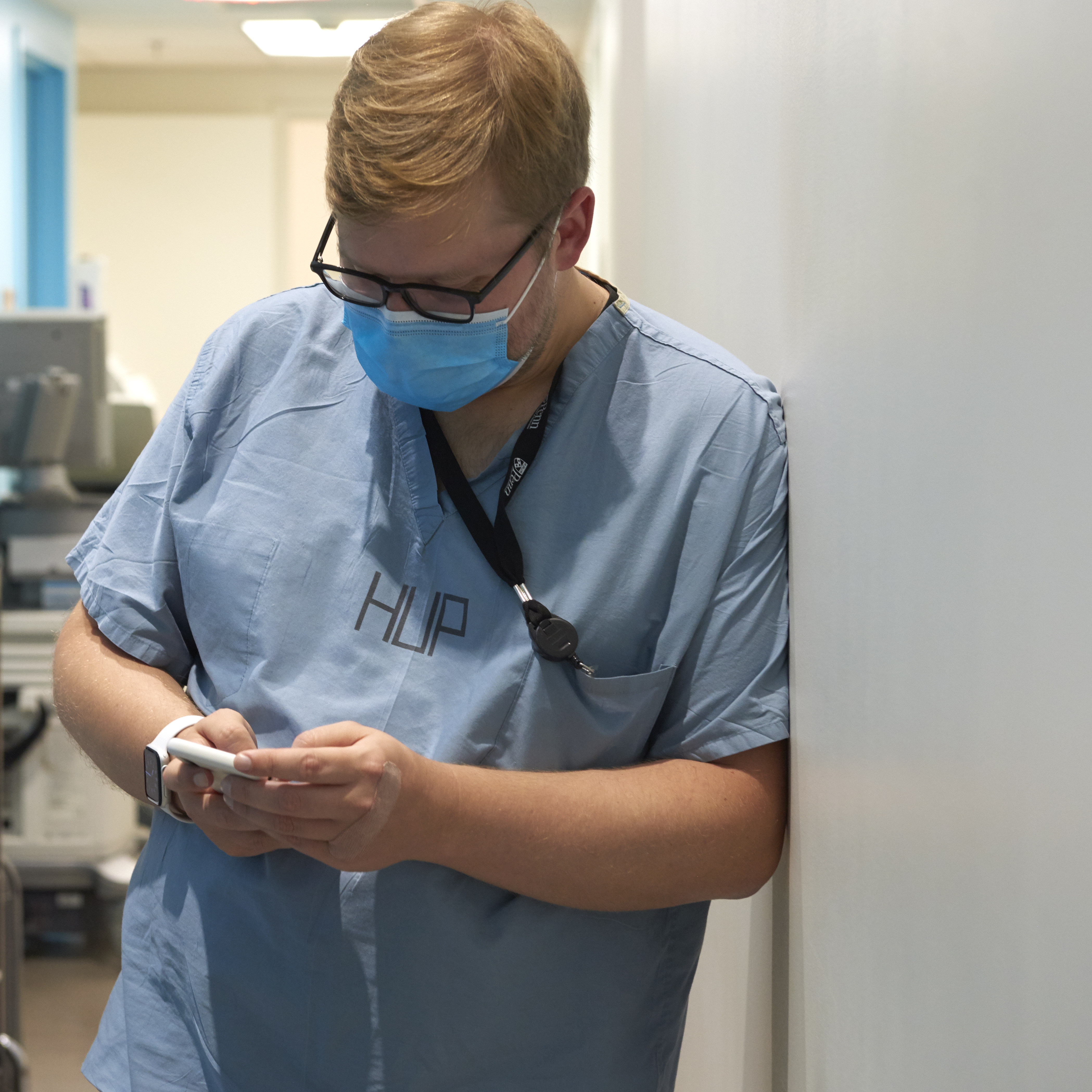 A male resident physician wearing HUP scrubs looks at his phone in a hallway during a break